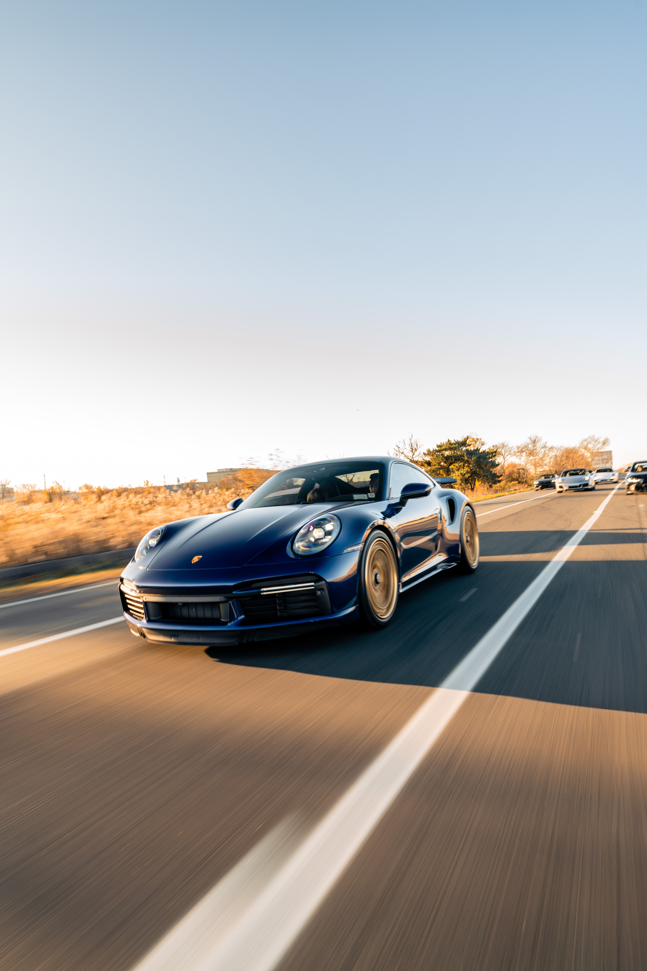A black sports car is driving on a highway at sunset, with other cars visible behind it, and trees and buildings in the background.
