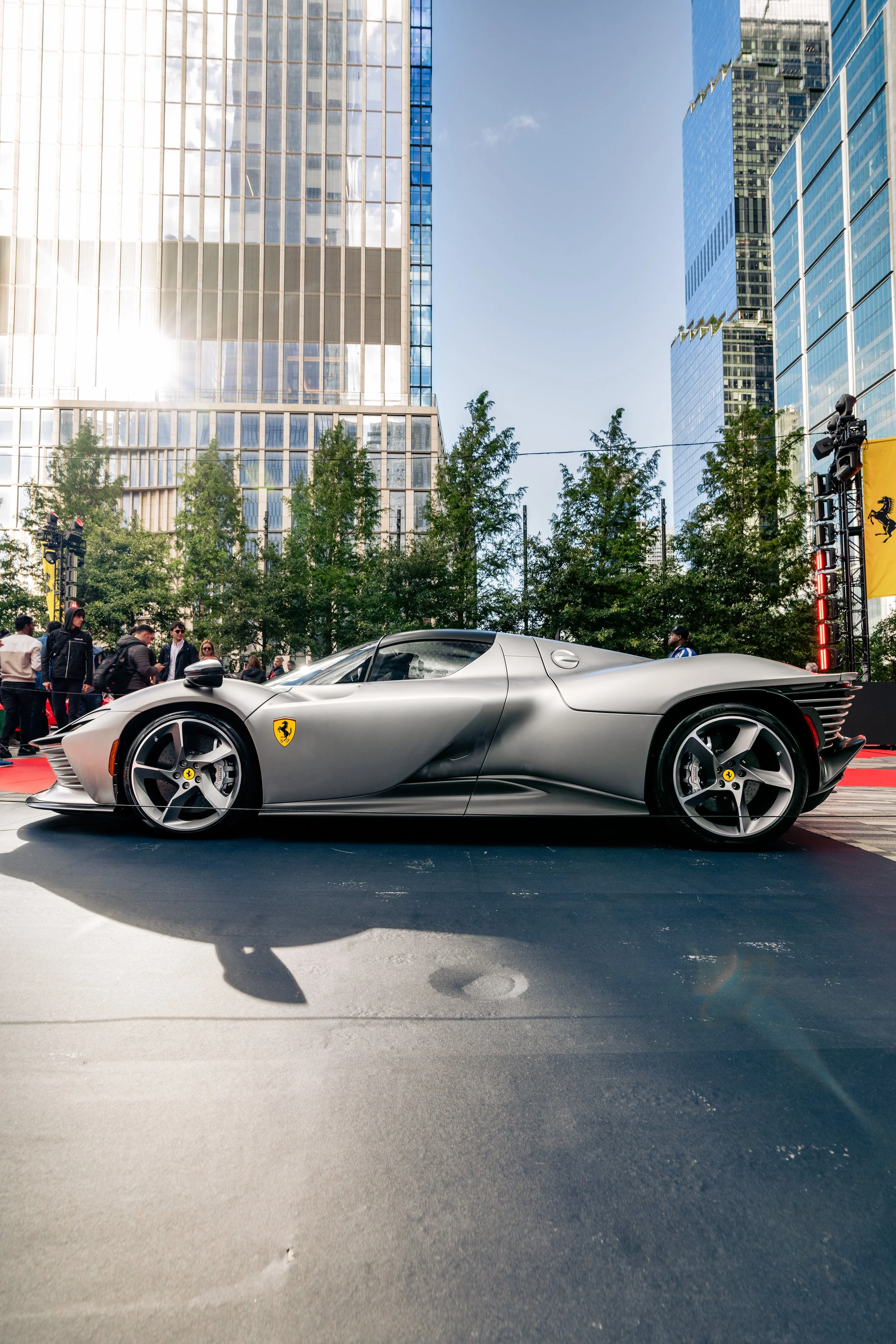 A silver Ferrari sports car parked on a city street with tall blue glass buildings and trees in the background. There are people walking nearby and a yellow Ferrari banner on the right side.