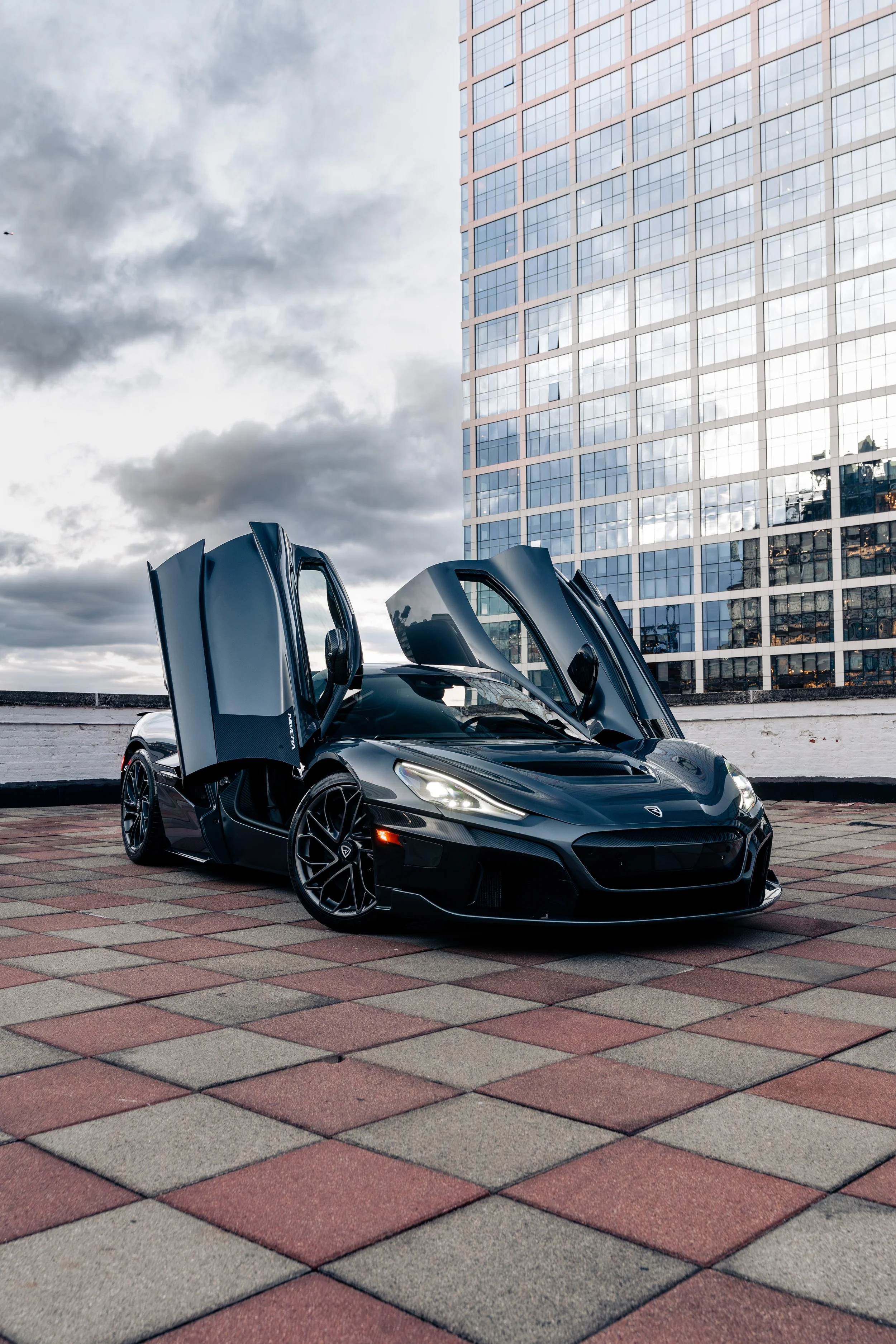 A black sports car with its doors open, parked on a tiled rooftop with a multi-story glass building and cloudy sky in the background.