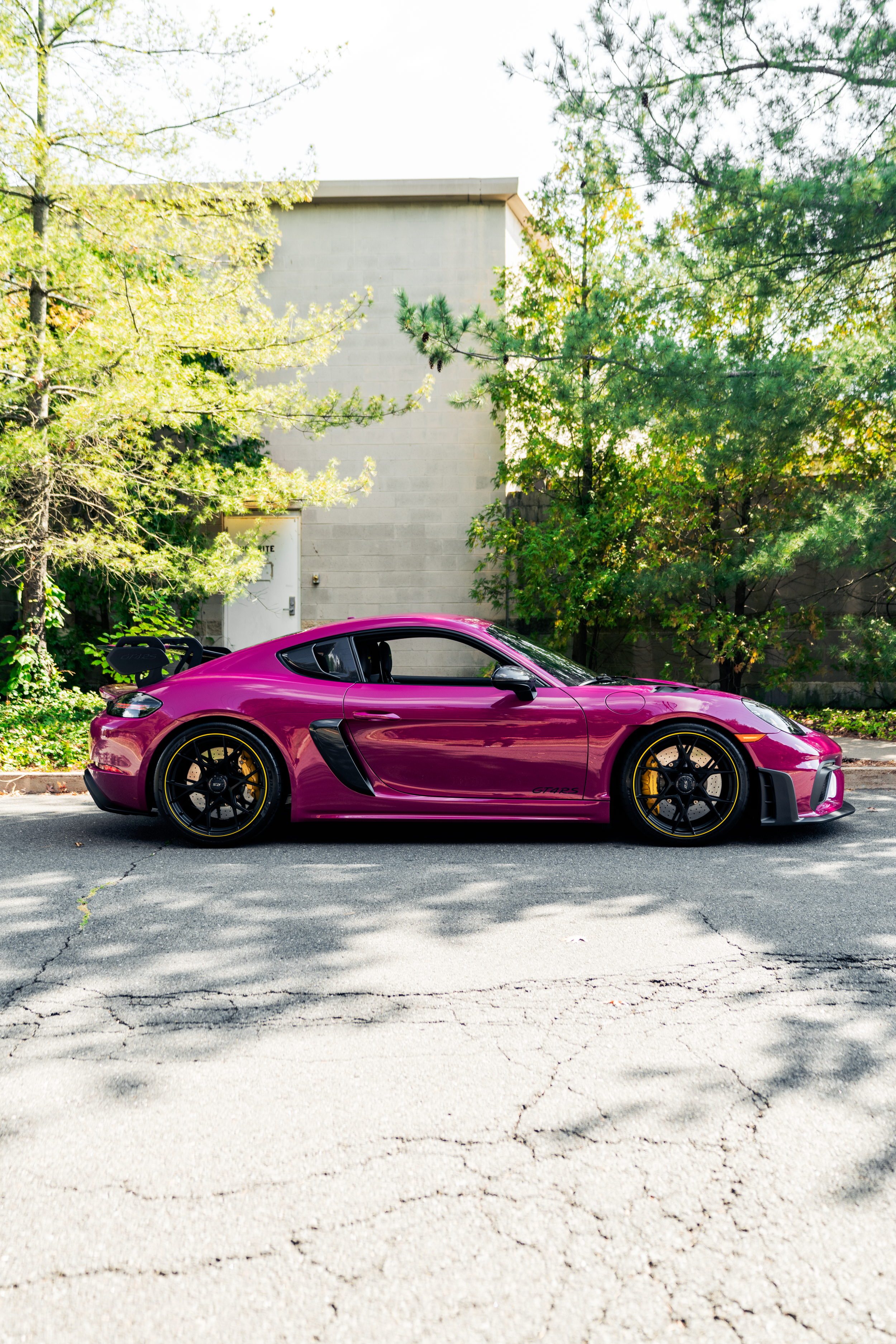 A bright pink sports car with black wheels parked on a street with cracked pavement. The background features green trees and a gray building wall.