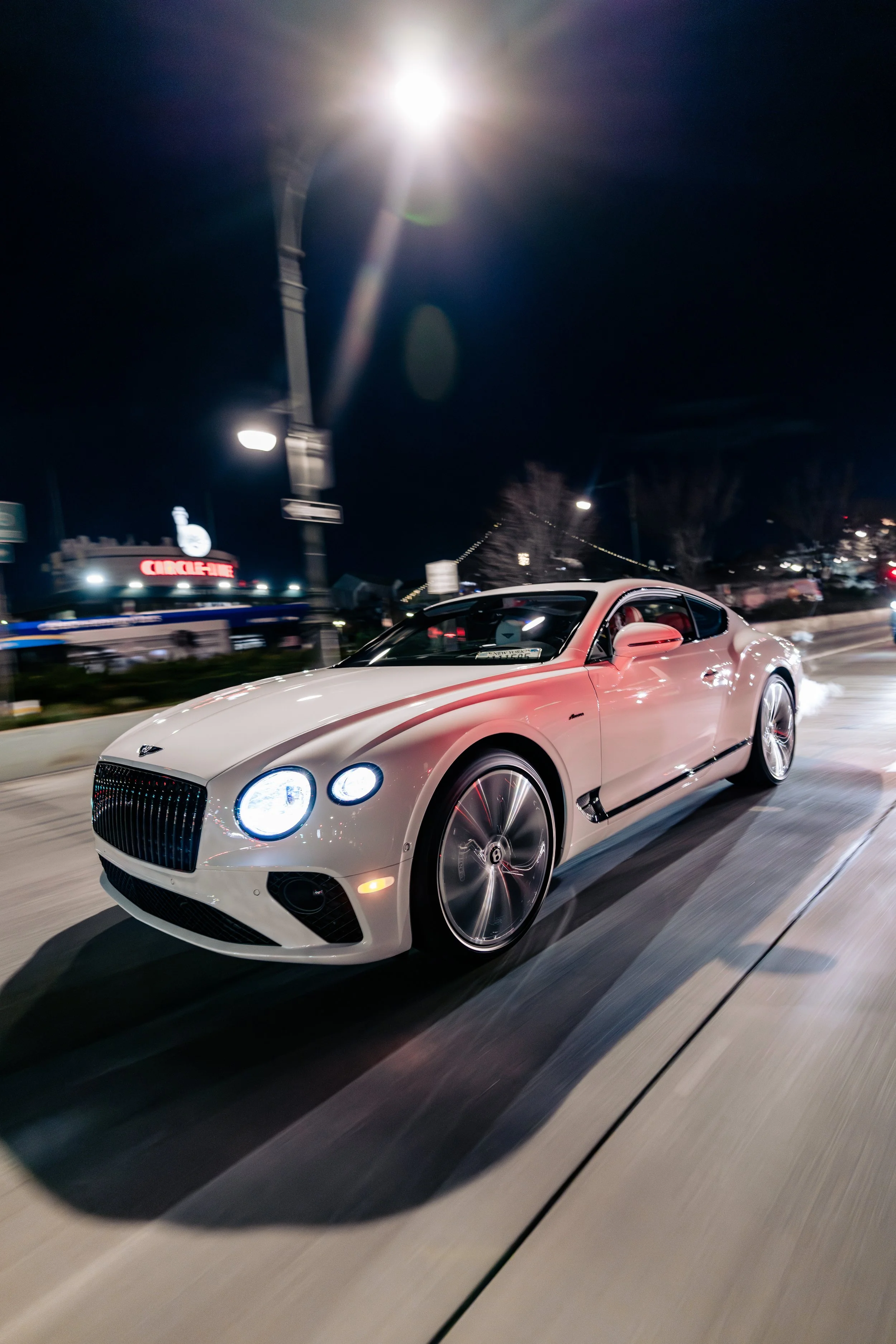 A white luxury sports car with black wheels and a black front grille driving on a city street at night, with bright streetlights and blurred background.