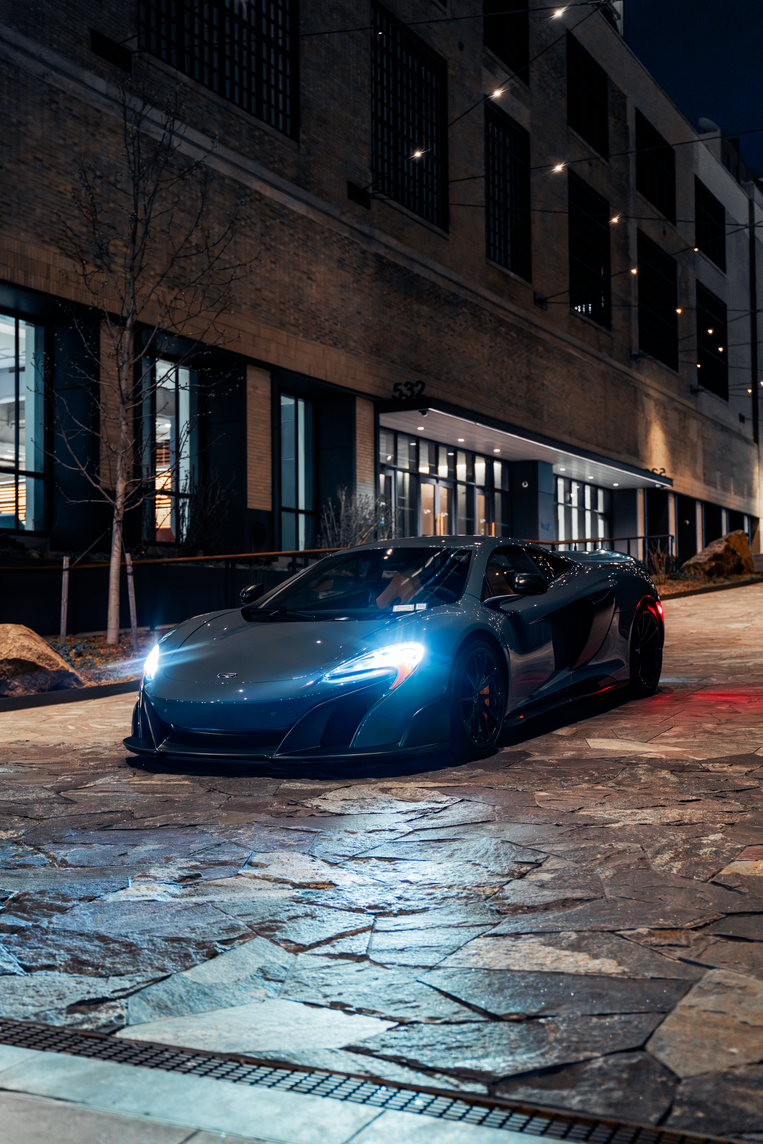 Luxury sports car parked on a street at night in front of a modern building with lit windows and string lights, with a leafless tree and rocky pavement in the foreground.