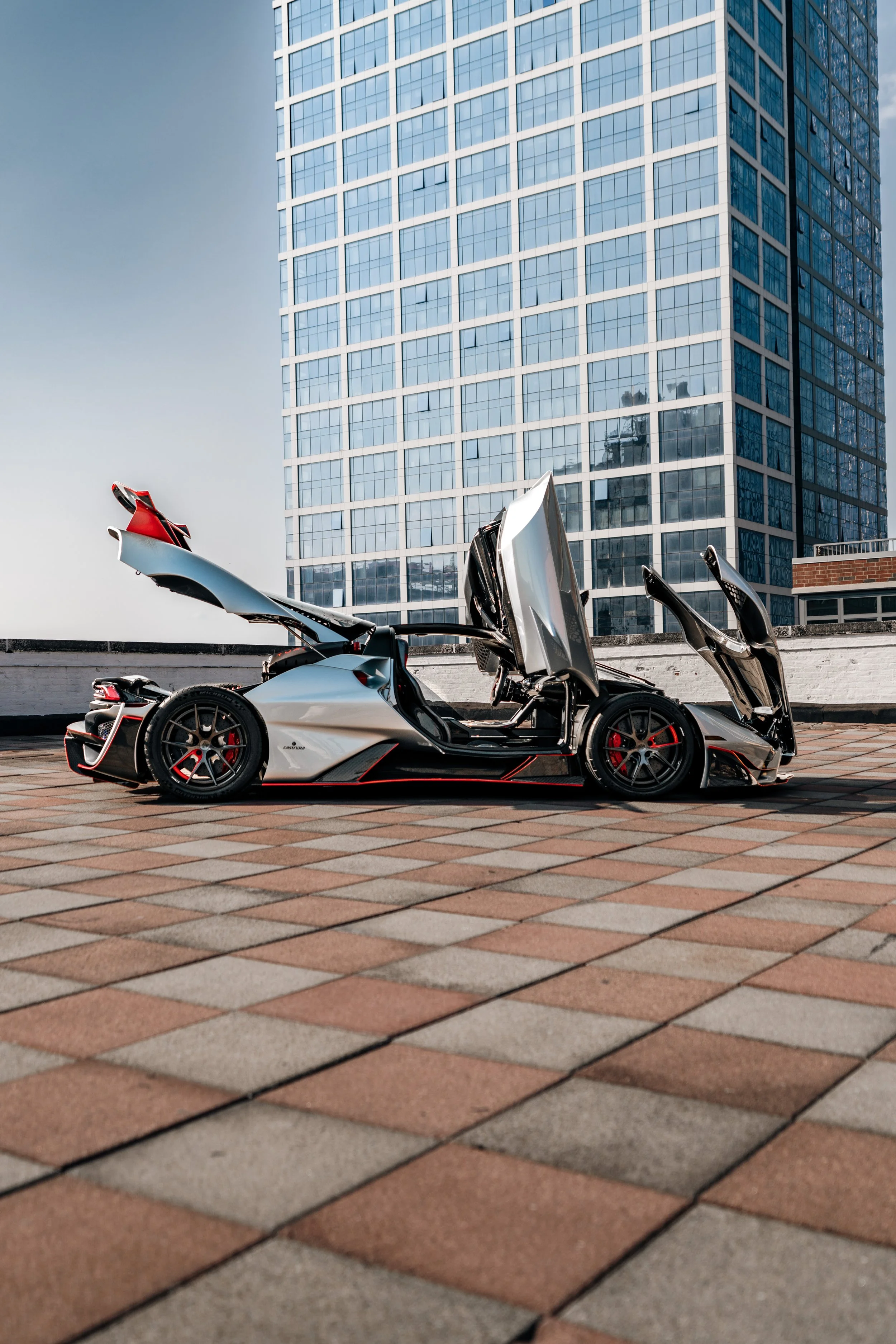 A white Lamborghini sports car with its doors open, parked on a rooftop with a brick patterned surface, in front of a tall modern glass building.