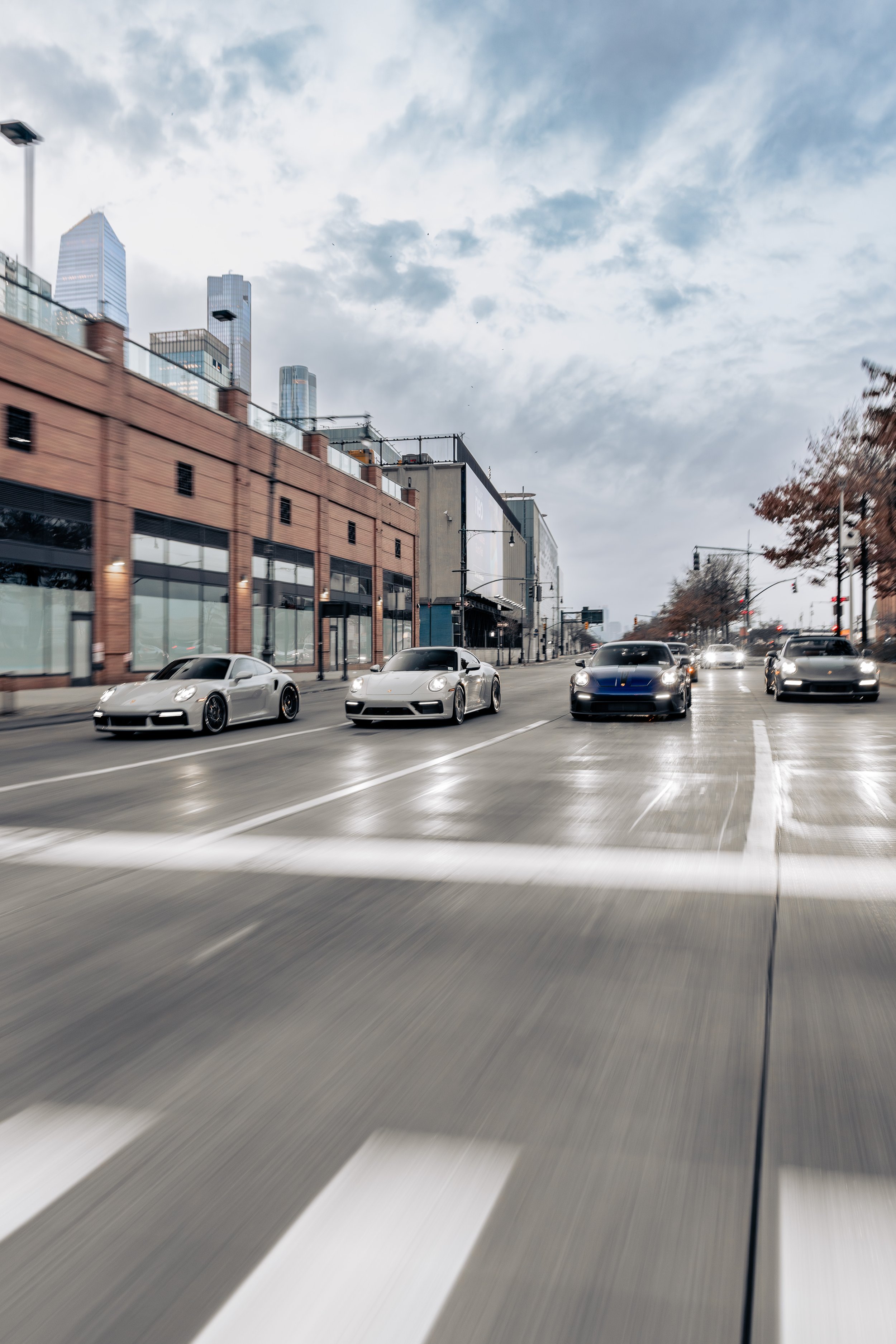 Street scene with multiple luxury sports cars driving on a city road, modern skyscrapers in the background, overcast sky.