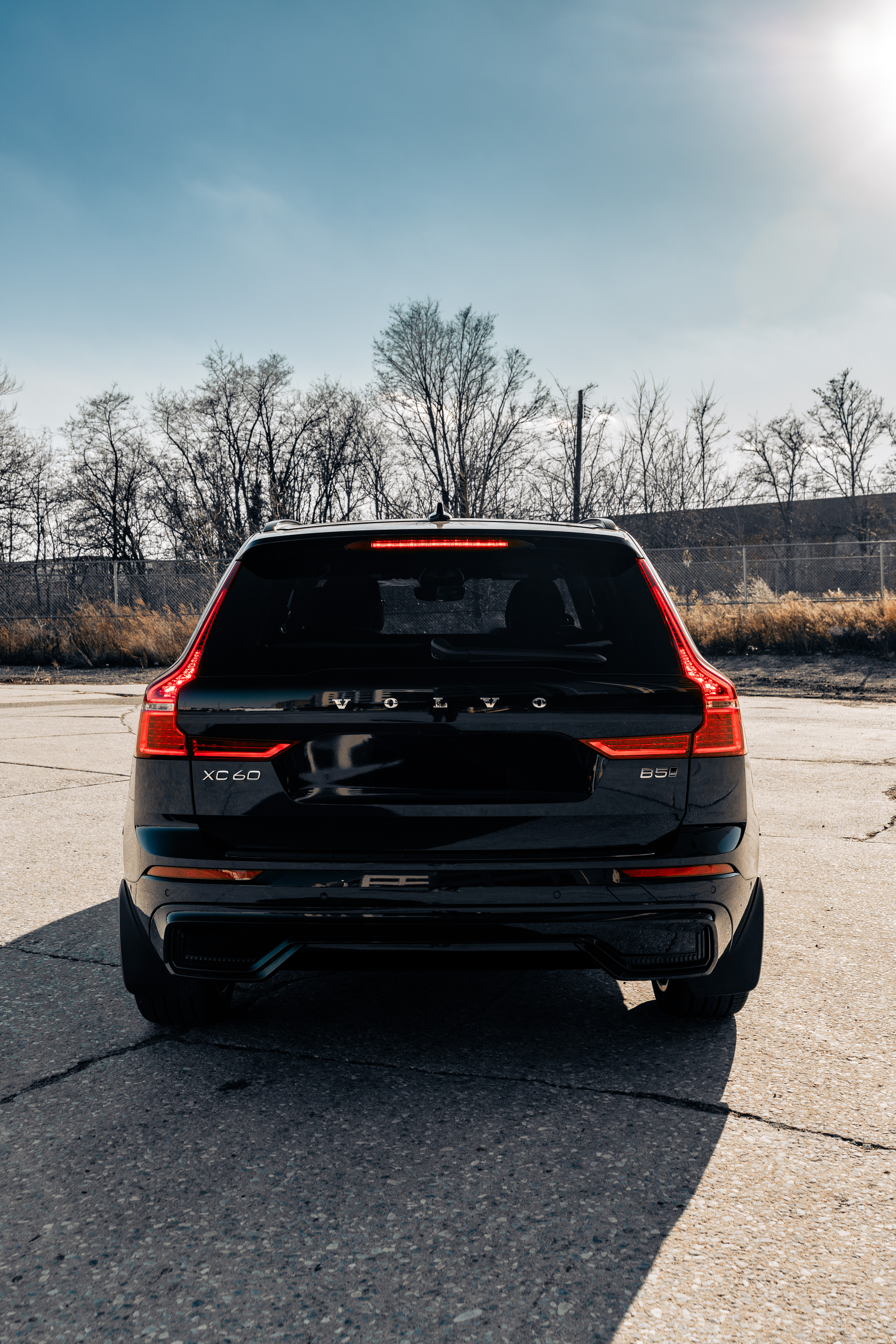 Rear view of a black Volvo XC60 B5 SUV parked outdoors on a paved surface, with leafless trees and a chain-link fence in the background, under a partly cloudy sky.