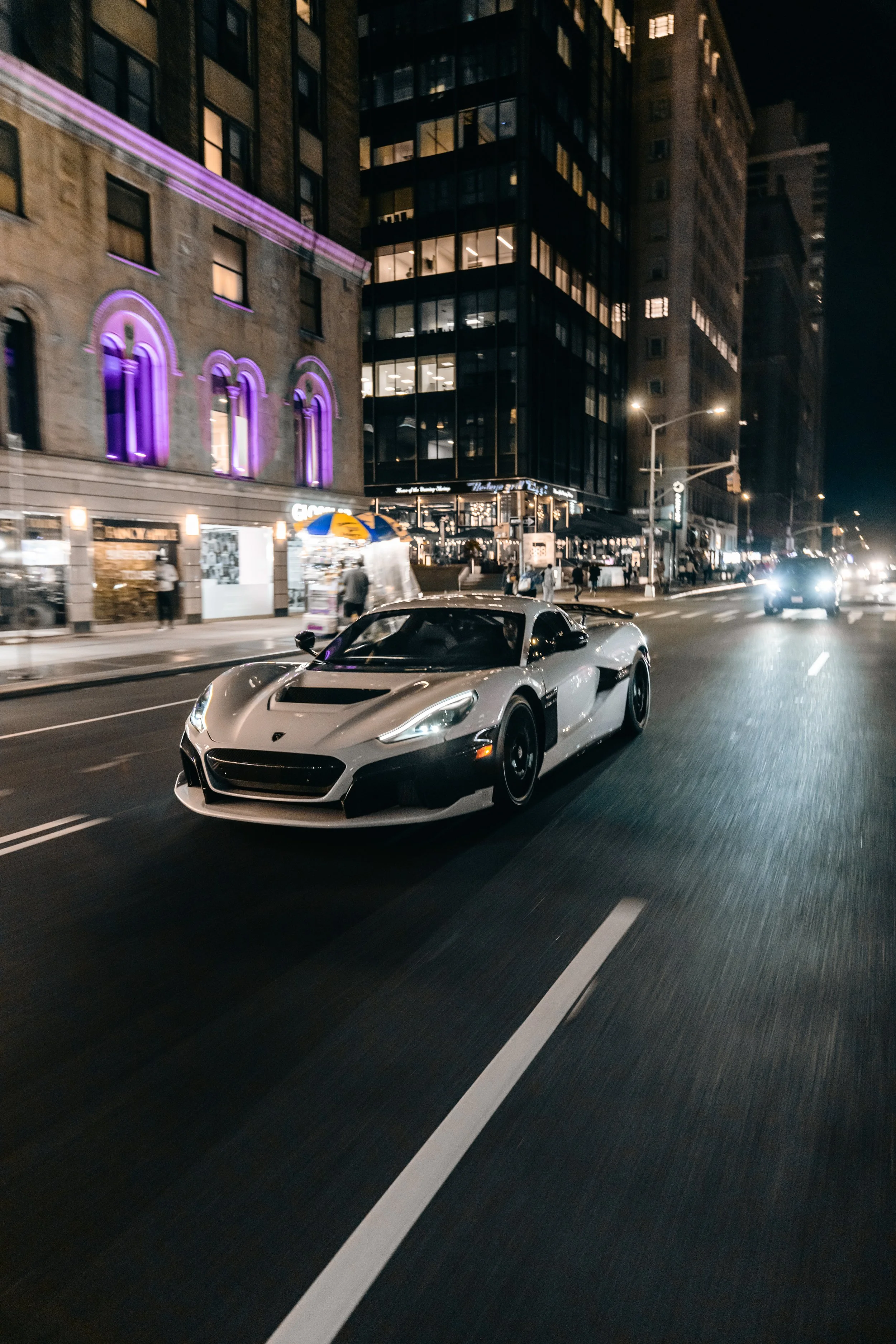 Nighttime city street scene with the Rimac Nevera driving in the foreground, tall buildings with lit windows, and a street vendor with a yellow and blue umbrella on the sidewalk.