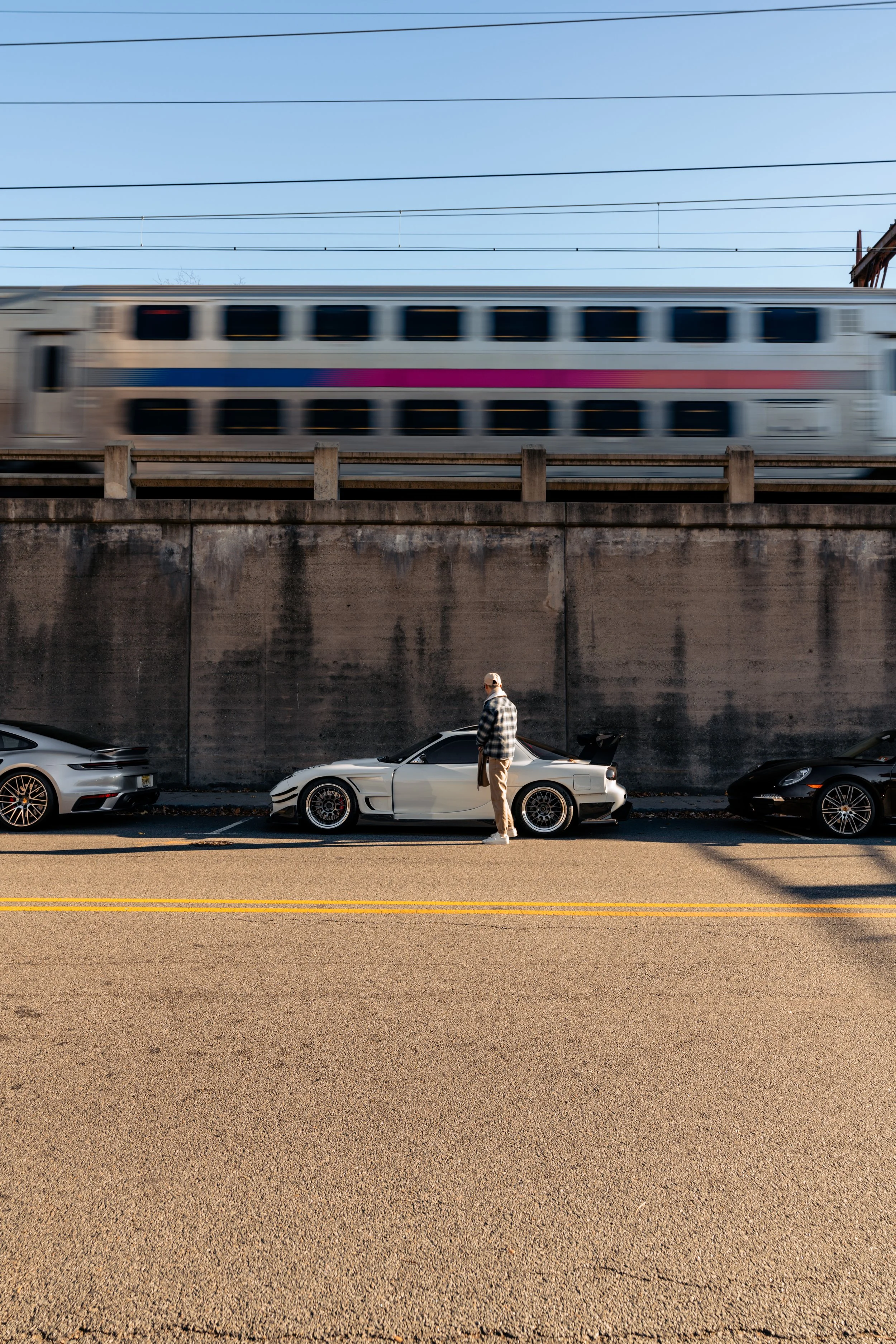 A man standing beside a white sports car on a street, with a concrete wall behind him. Above, a moving train passes on elevated tracks, and a blue sky with power lines is visible.