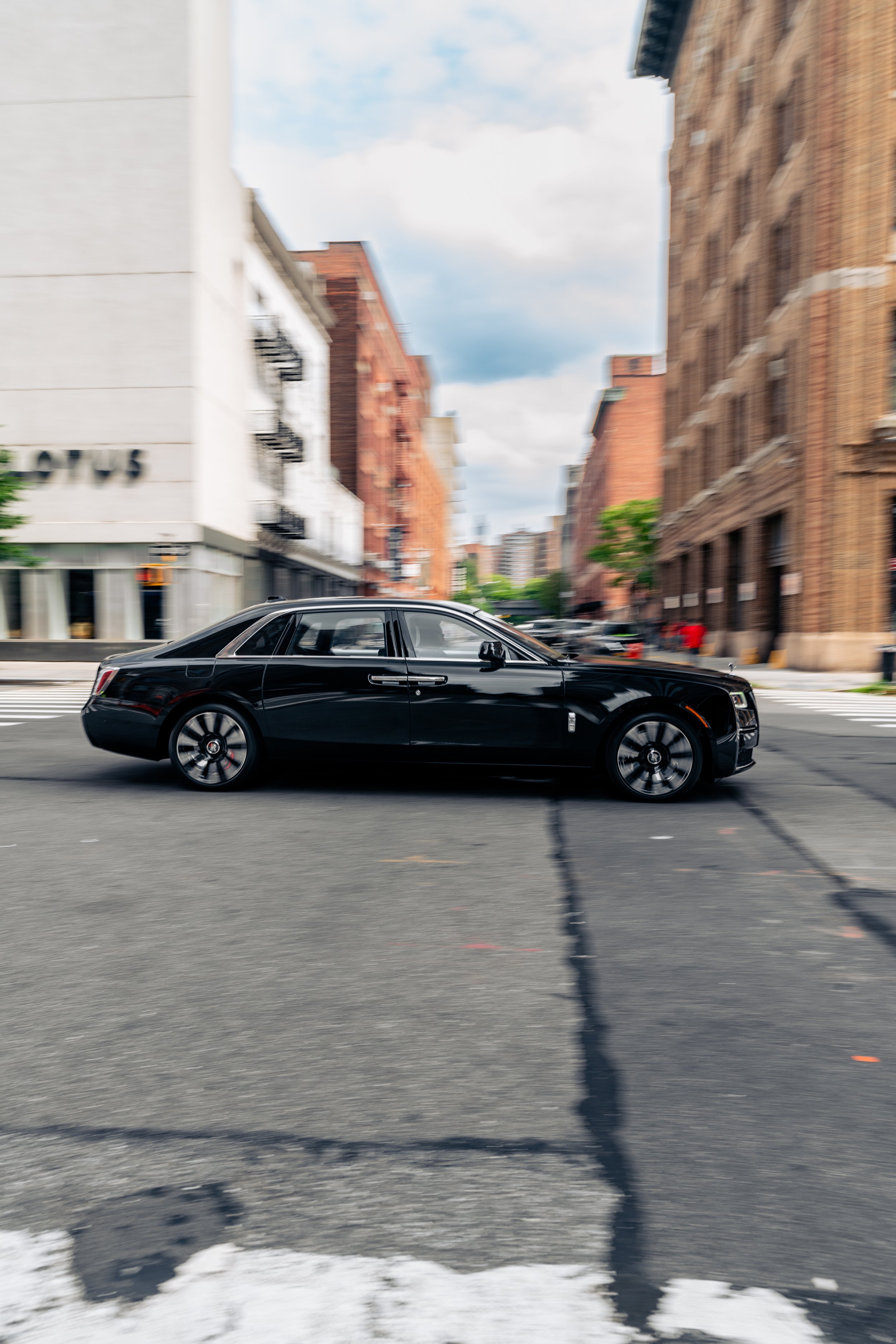 A Rolls Royce Ghost driving on a city street with brick and modern buildings in the background.