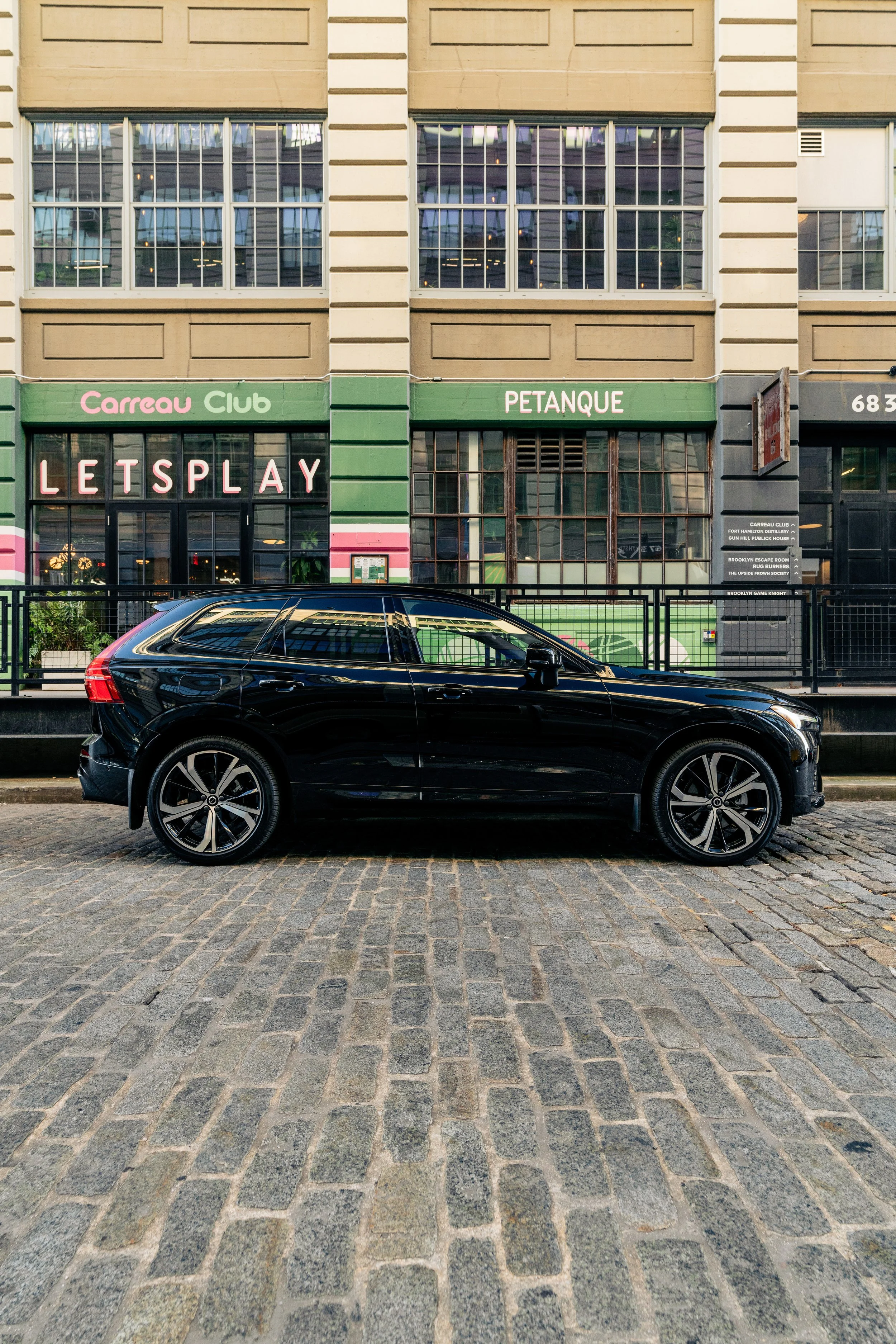 A black car parked on a cobblestone street in front of a building with large windows and green storefronts, displaying signs for Carreau Club, Petanque, and LetsPlay.