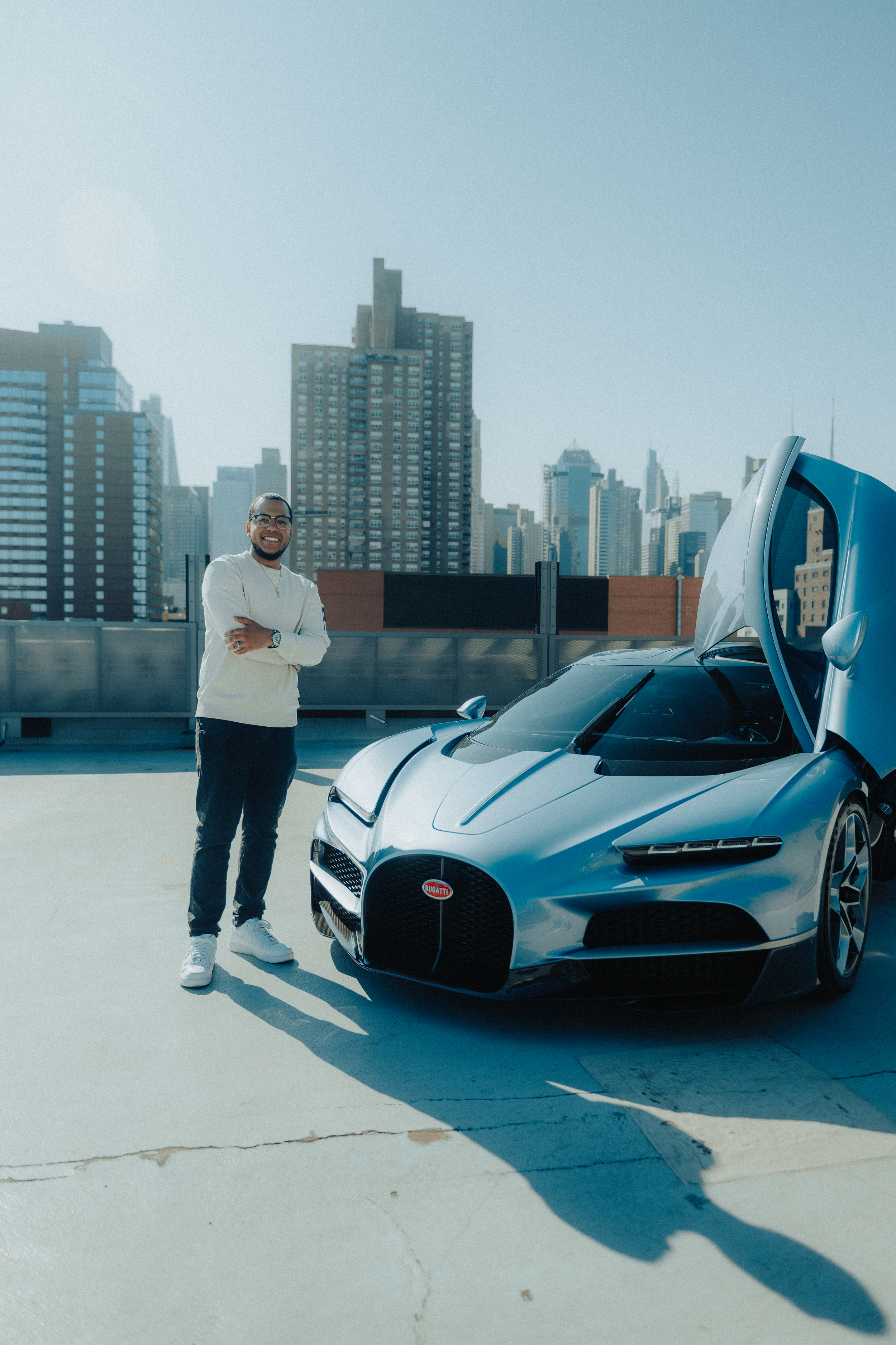 A man standing next to a silver Bugatti sports car with his arms crossed, on a rooftop parking lot with a city skyline in the background, under a clear blue sky.