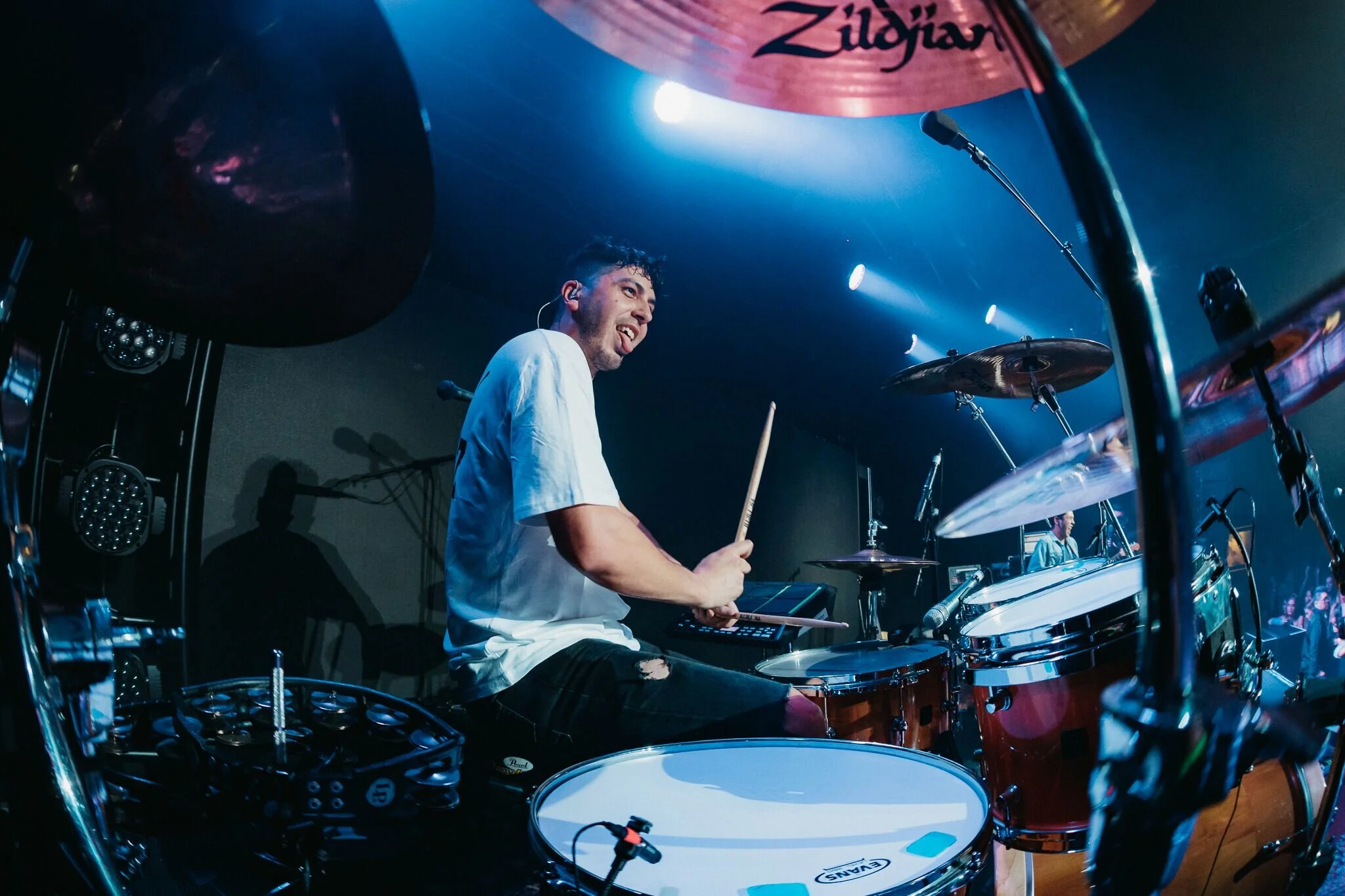 Drummer playing a drum kit on stage at a live concert, surrounded by cymbals and stage lighting.