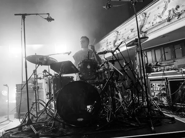 Black and white photo of a drummer playing a drum set on stage with overhead microphones and lighting equipment.