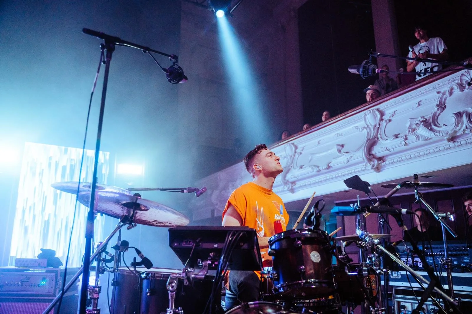 Drummer performing on stage, surrounded by drum kit and equipment, with audience in a balcony.