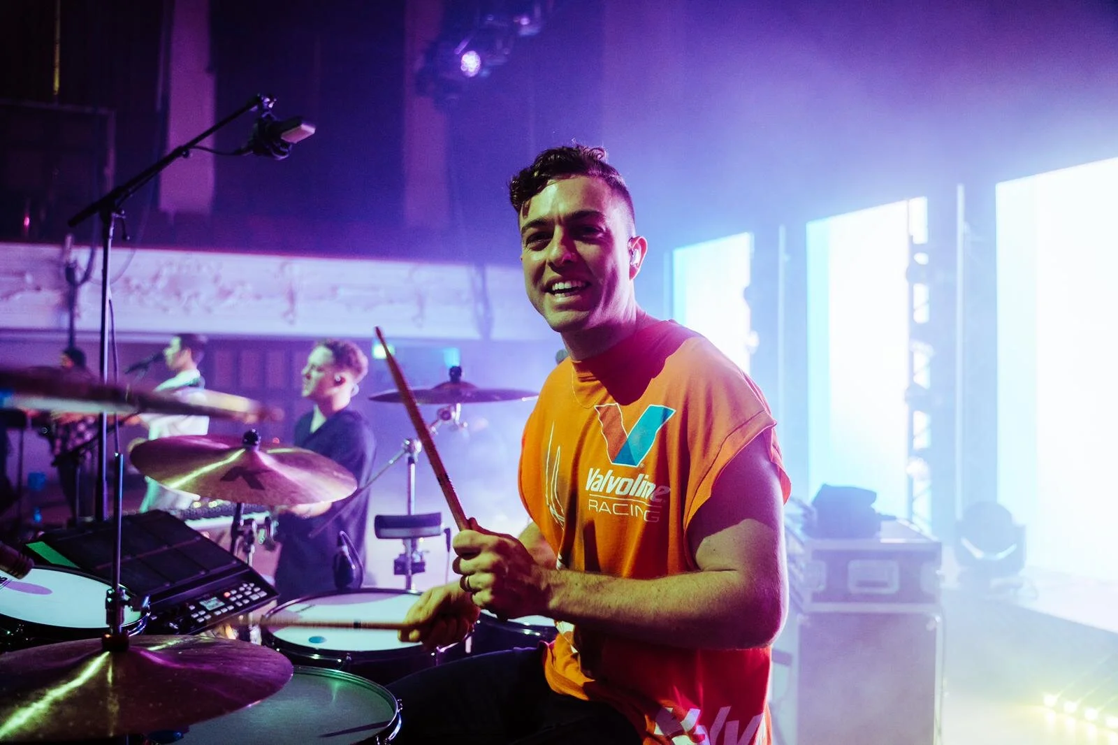 Drummer playing on stage with orange Valvoline shirt, smiling and holding drumsticks. Musical equipment and other band members in the background.