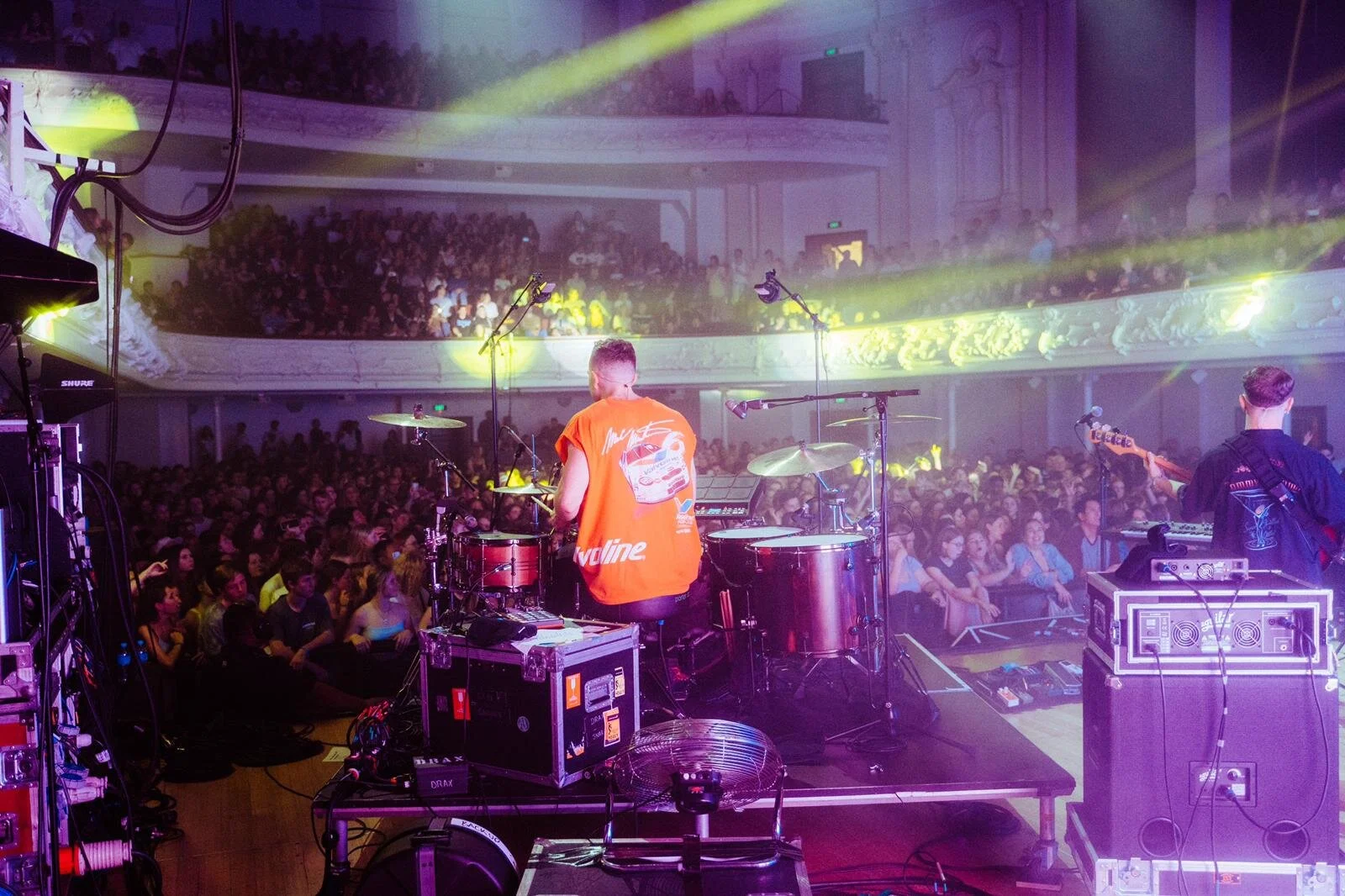 Live concert scene with band performing on stage, drummer in red shirt, audience in packed theater, bright colorful lights.