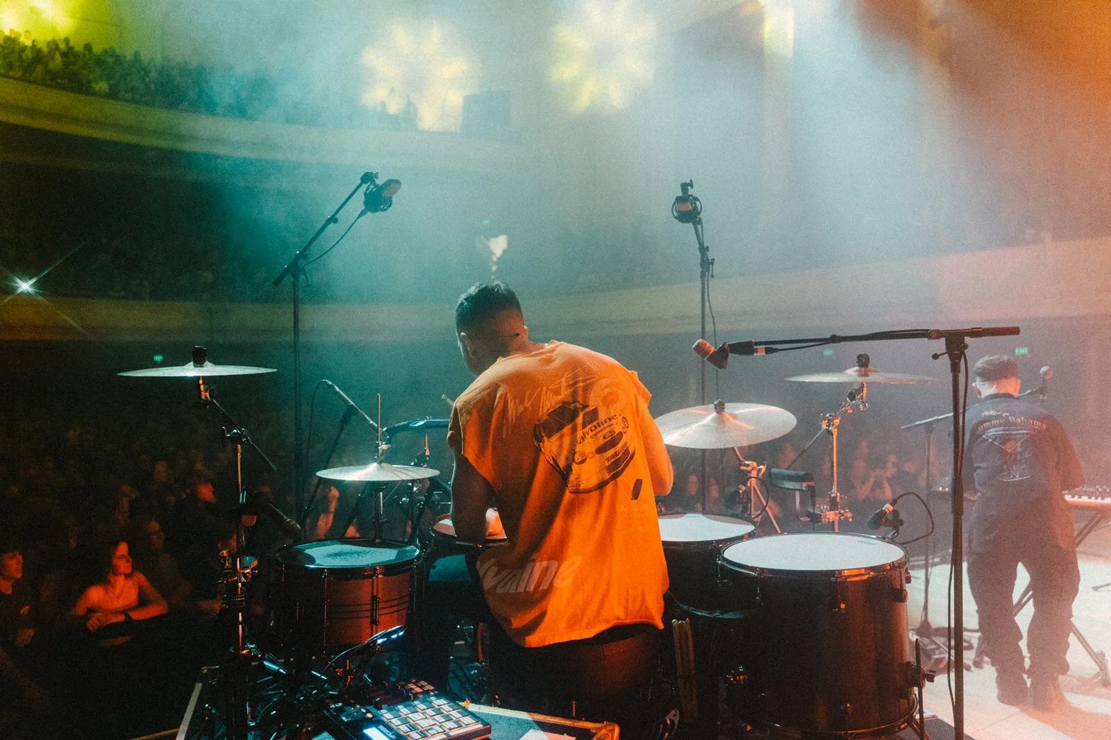 Drummer and musician performing on stage in front of a large audience, with drum set and microphones visible under colorful lighting.