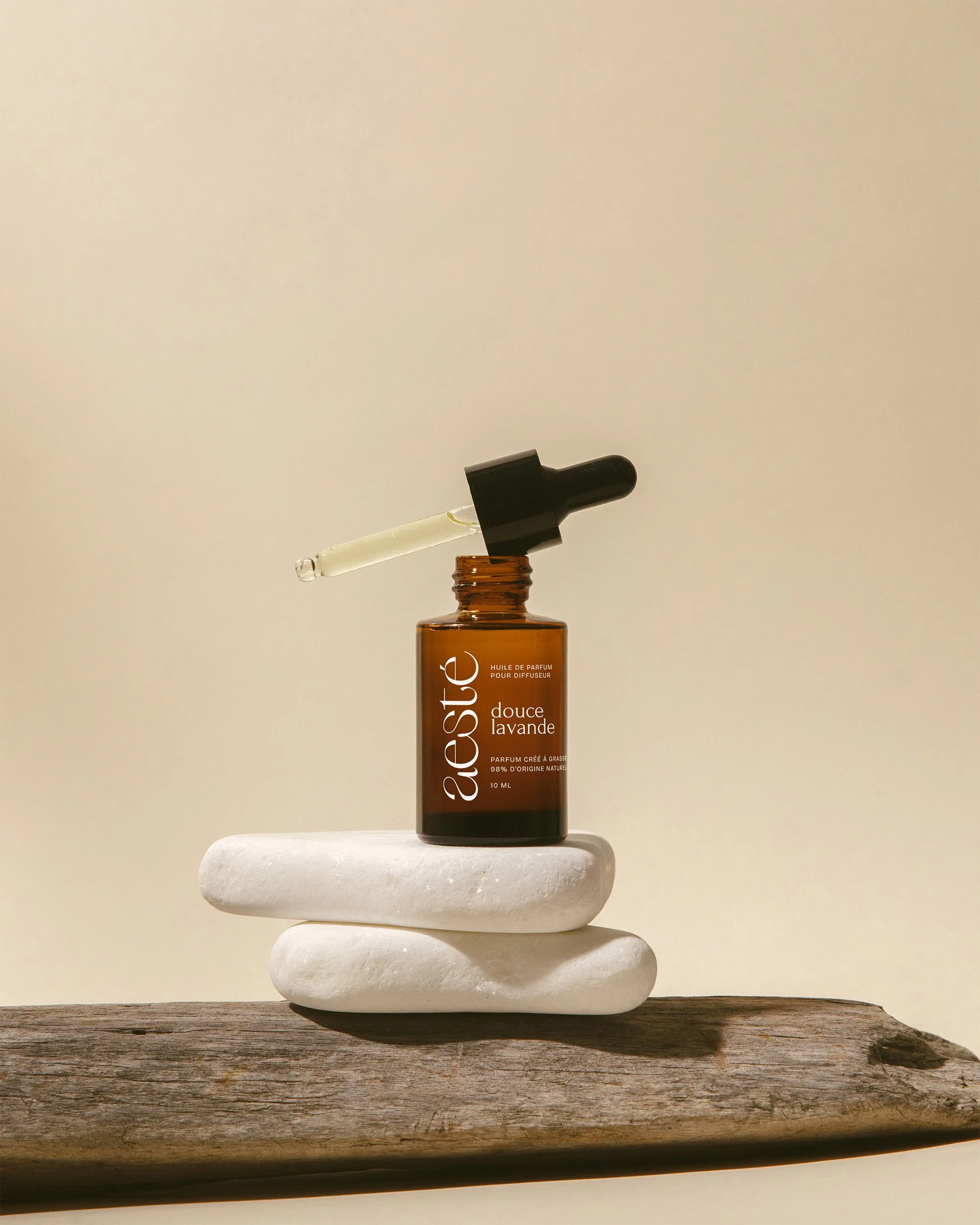 Une bouteille d'huile de parfum ambrée avec un compte-gouttes noir posé sur deux galets blancs, sur une planche en bois, fond beige