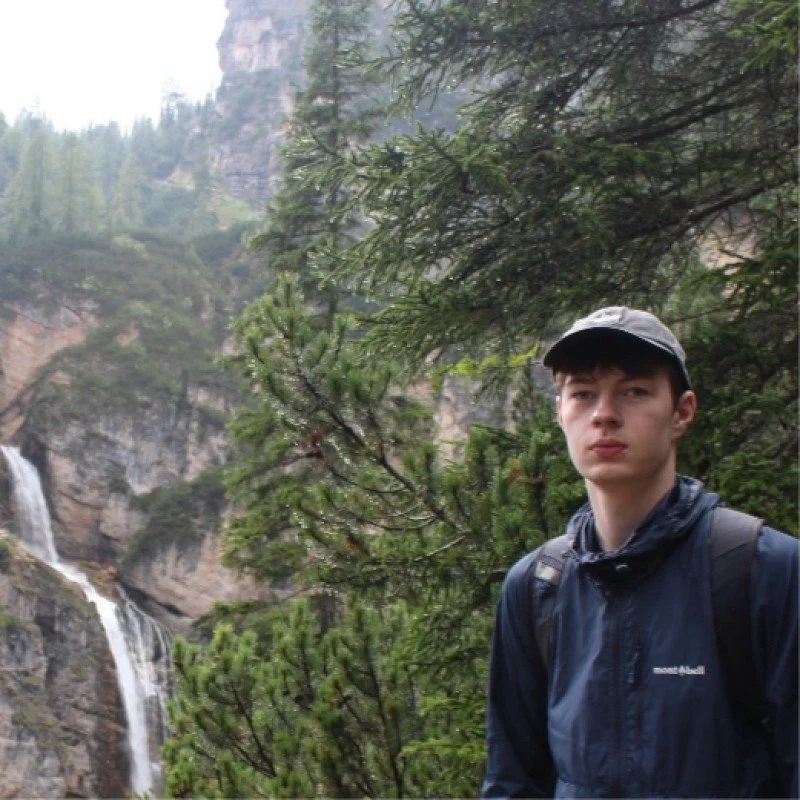 A man with curly dark hair, glasses, a nose ring, and a beard, looking off to the side with a slight smile.