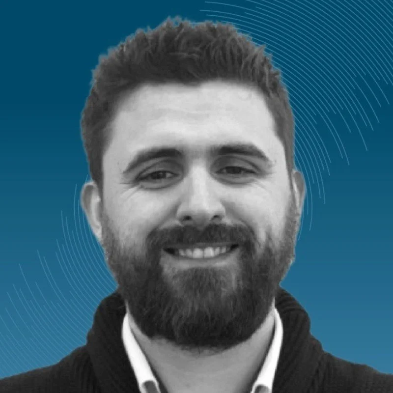 A smiling woman with curly black hair, wearing glasses, hoop earrings, layered necklaces, and a white top with colorful stripes.