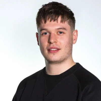A young woman with shoulder-length black hair smiling outdoors with greenery in the background.