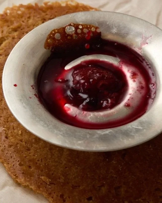 Close-up of a silver bowl containing dark red berry jam or syrup, with a spoon partially submerged, resting on a brown, crispy bread or cake.