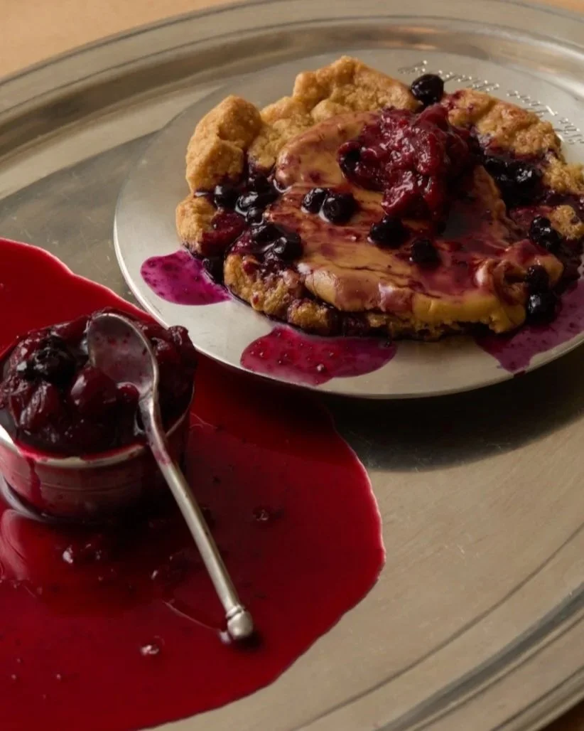 a galette with berries on a silver tray, beside a cup of compote spilling across it.