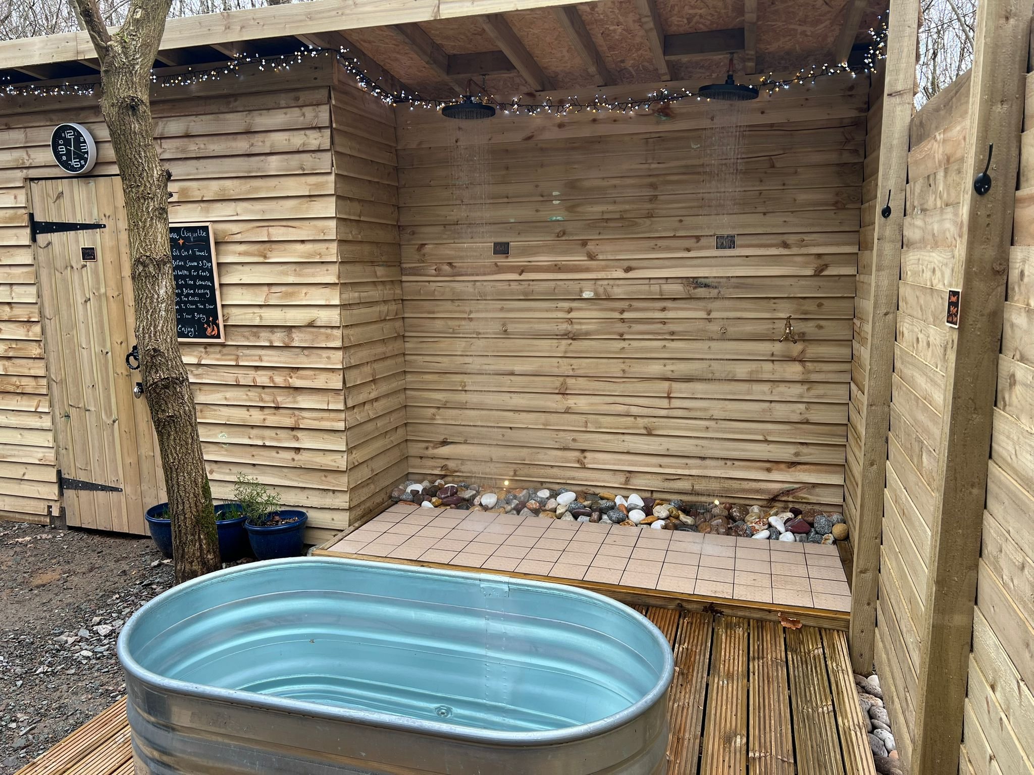 An outdoor shower area with a wooden privacy wall and ceiling, a small tiled platform, a rock bed, two potted plants, a metal soaking tub, and a chalkboard sign.