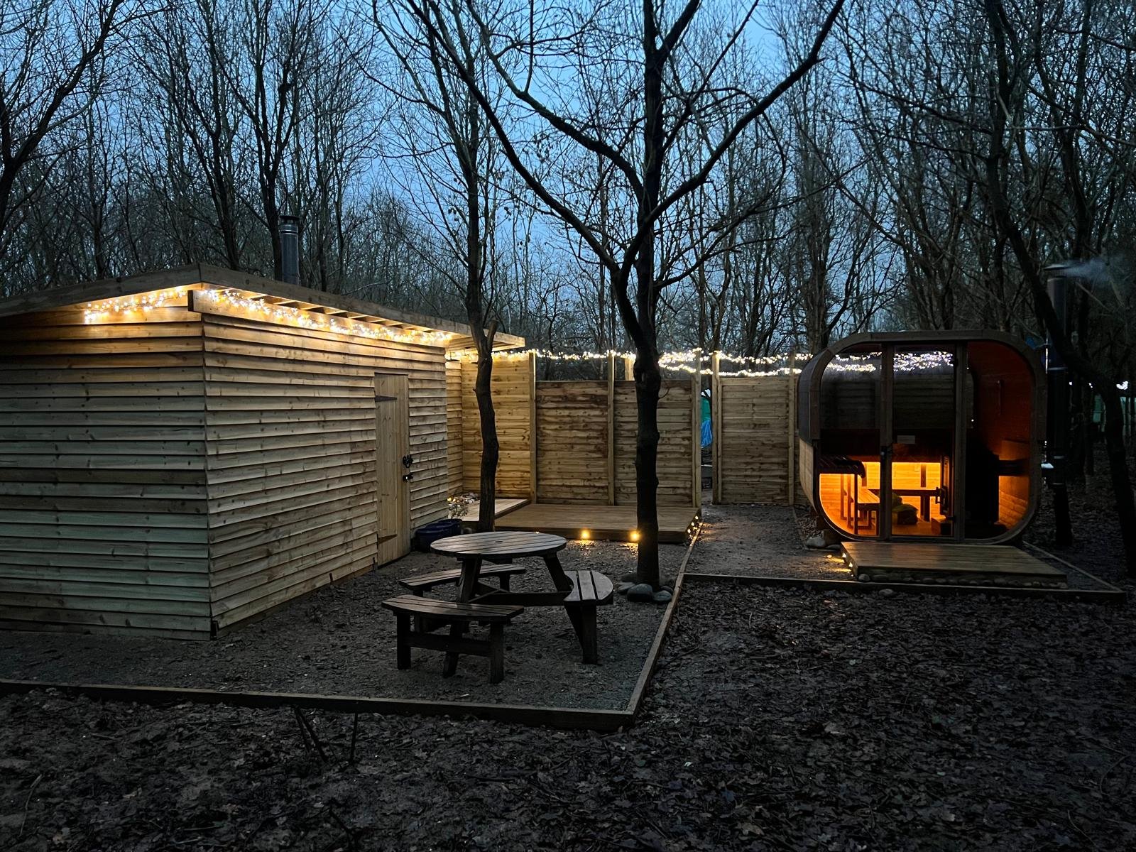 A cozy glamping site in a wooded area at dusk, featuring a wooden cabin with string lights, a small outdoor seating area with a picnic table, and a modern barrel-shaped tiny home with warm interior lighting.