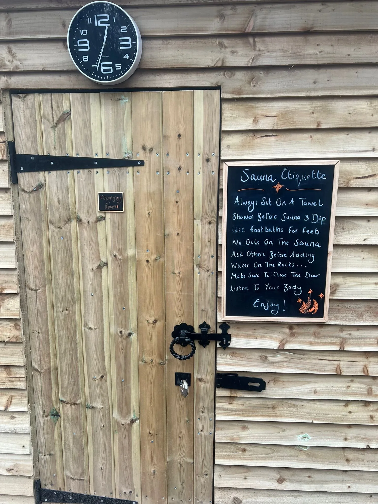 A wooden door with a sliding barn door lock. A clock showing 12:56 is mounted on a wooden wall above the door. To the right of the door, there is a blackboard with instructions about sauna etiquette, including sitting on a towel, showering before entering, using foot baths, no oils on the sauna, and listening to your body, with decorative drawings of stars and a fox.