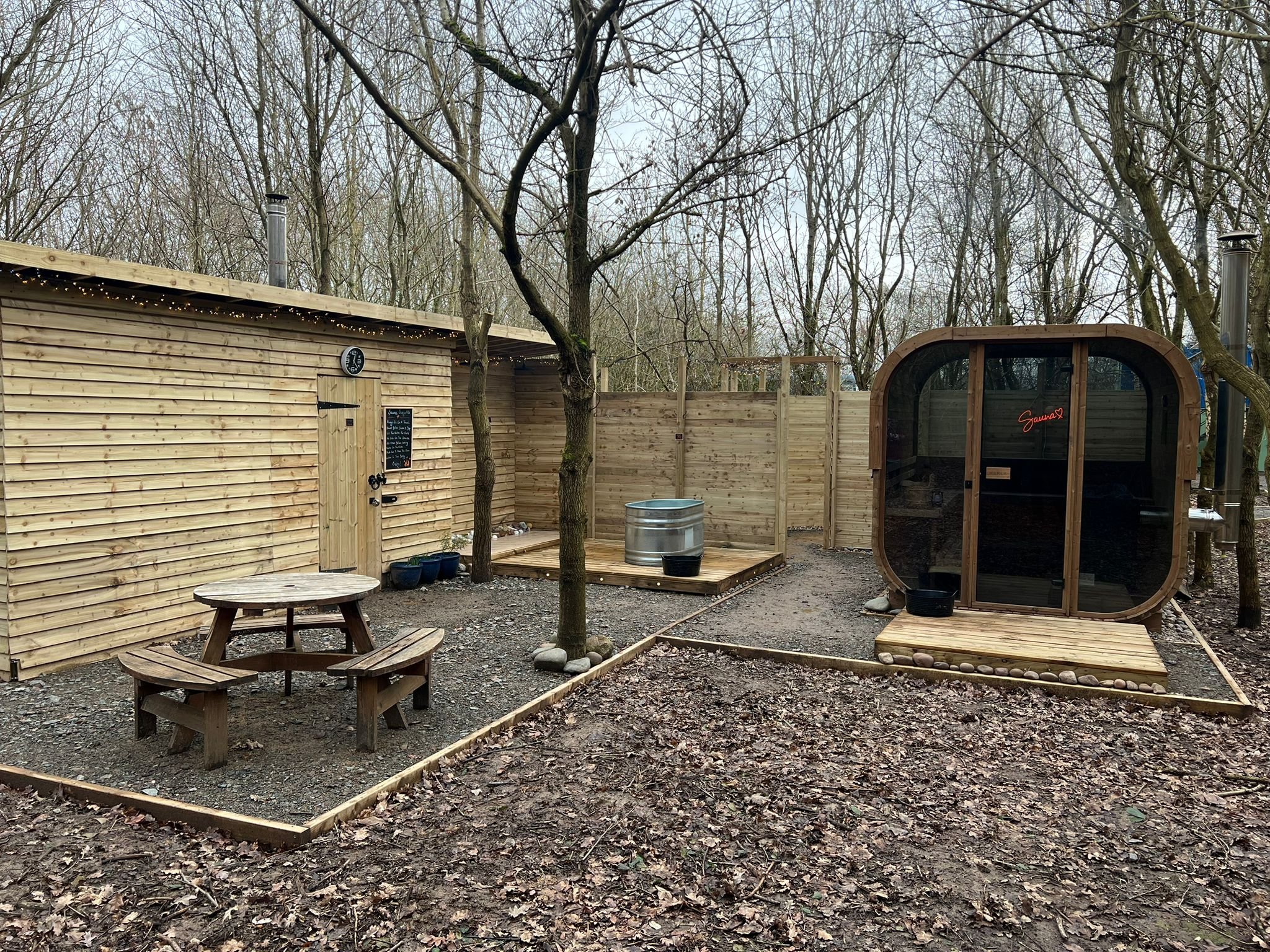 A woodland sauna, a small deck with a metal cold plunge tub, and a wooden picnic table with benches, surrounded by trees.