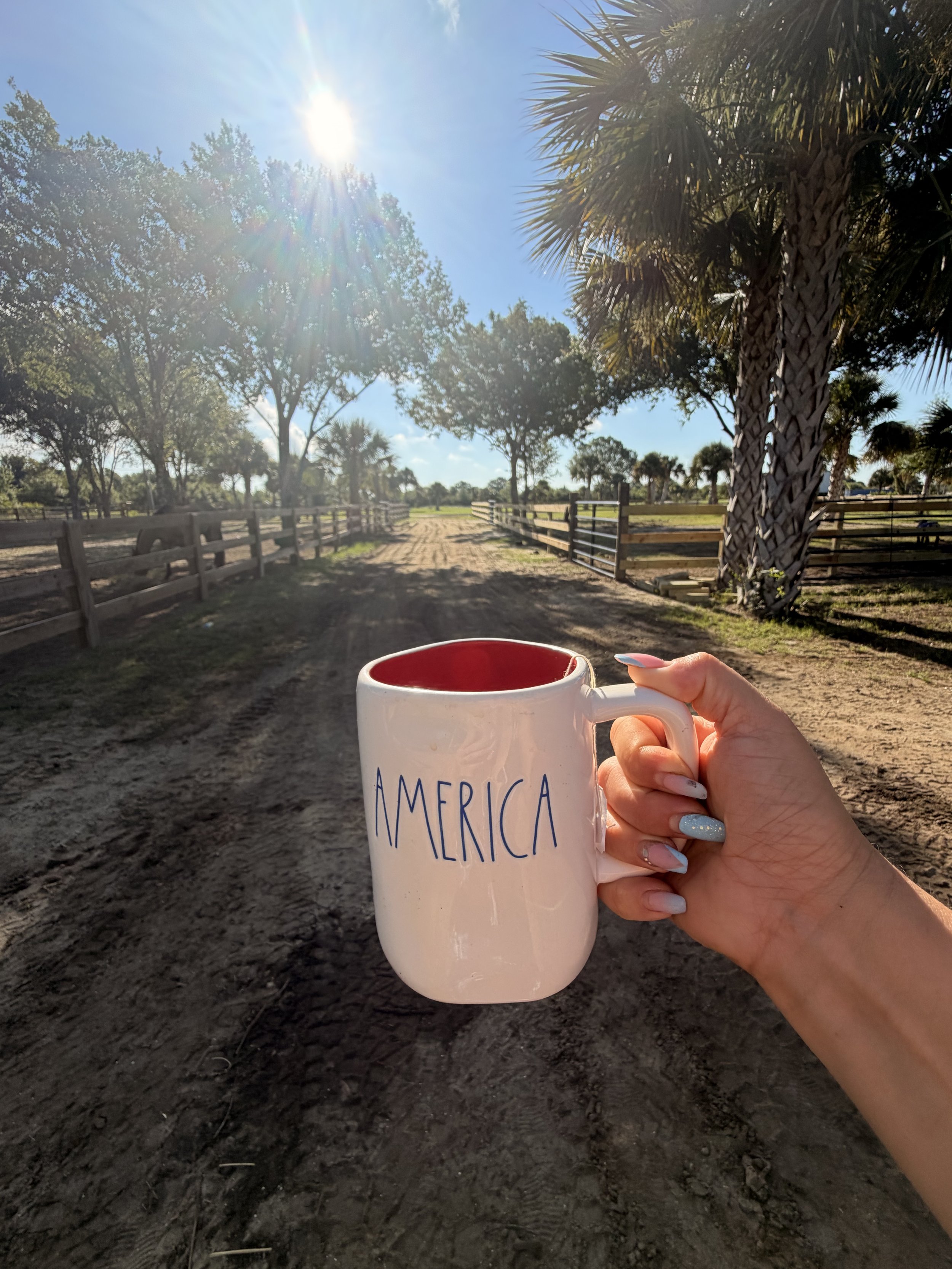 A person holding a white mug with the word "America" written on it, outdoors in a sunny area with trees, a dirt path, and a wooden fence.