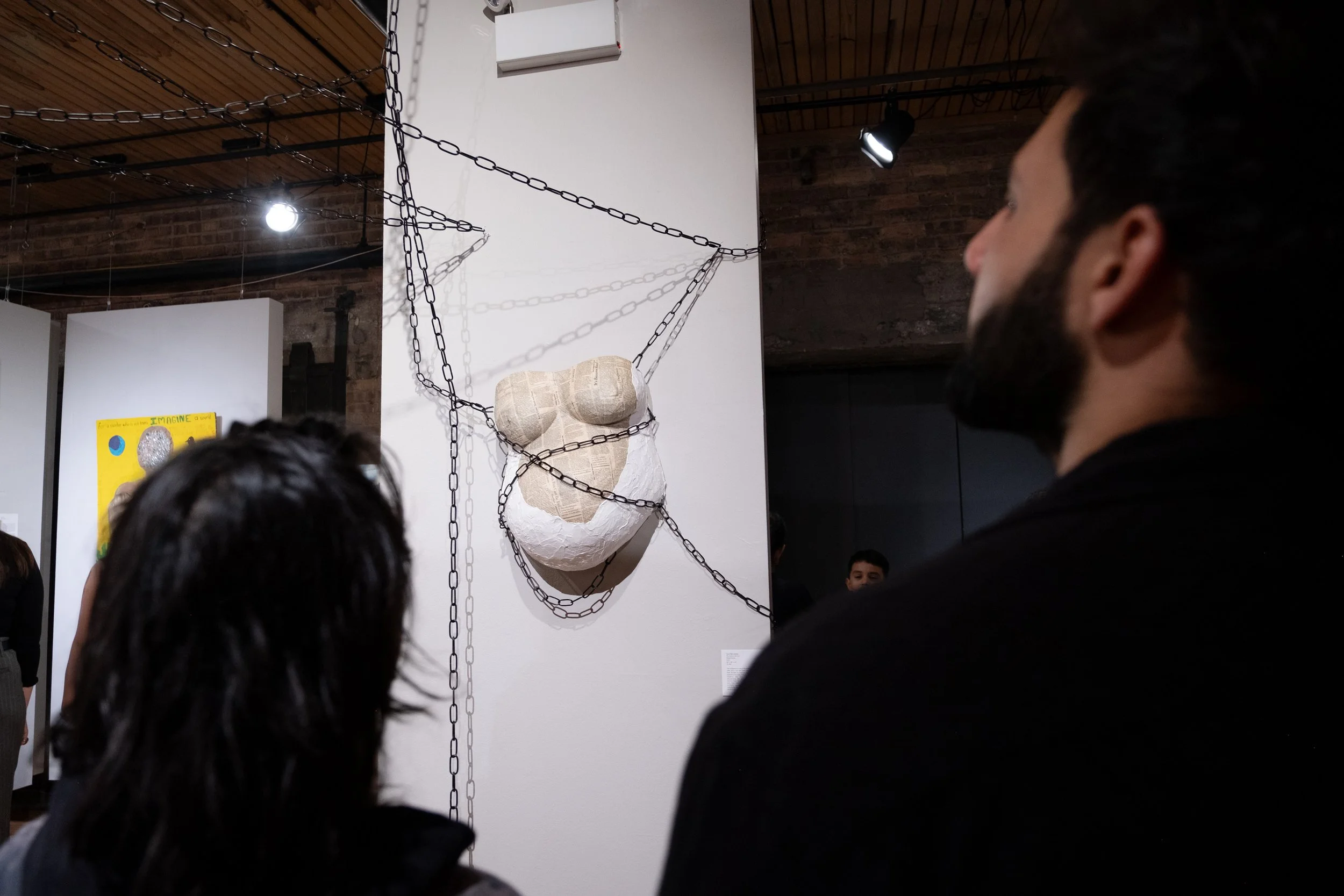 People viewing a mixed-media art piece resembling a face, mounted on a white wall with hanging chains, in an art gallery setting.