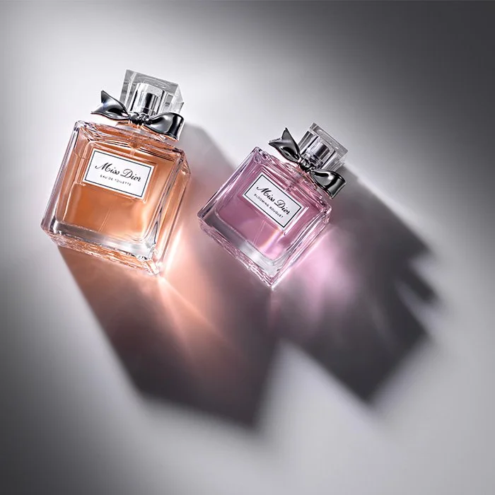 Two Miss Dior perfume bottles, one in peach and one in pink, with black bows on the caps, casting shadows on a light and dark background.