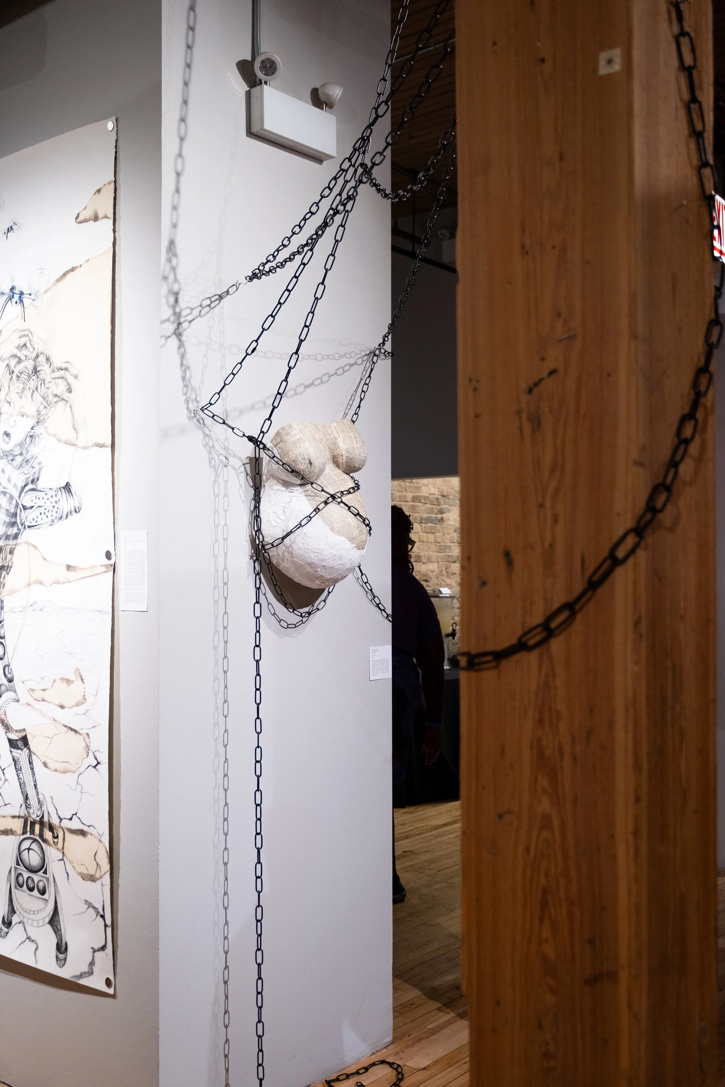 An art installation featuring chains wrapped around a 3D mask sculpture, displayed on a white wall with a drawing of a horse and a person to the side, in an indoor gallery setting.