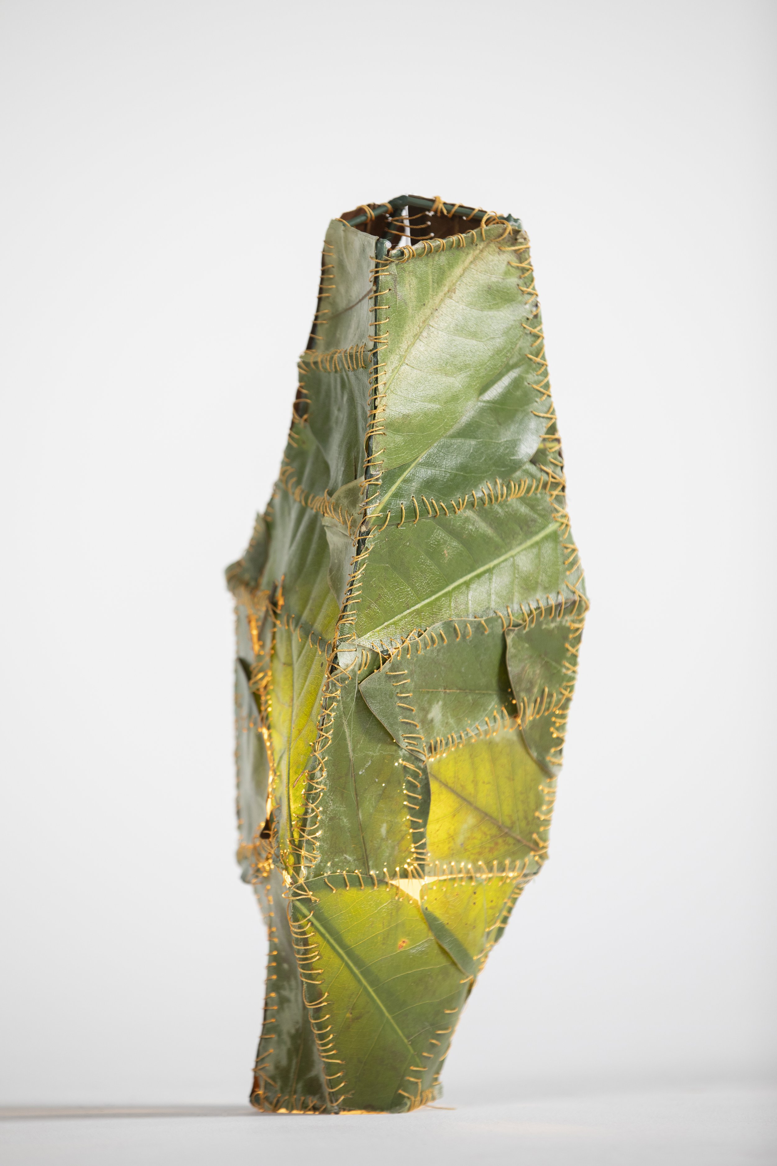 An art sculpture made of leaves and branches stitched together with orange string, shaped like a tall, narrow obelisk.