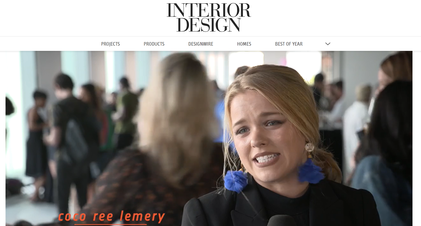 A woman with blonde hair tied in puffs with blue hair ties, wearing earrings and a black blazer, speaking in an interview. Behind her, there are people gathered in a bright indoor space with large windows.
