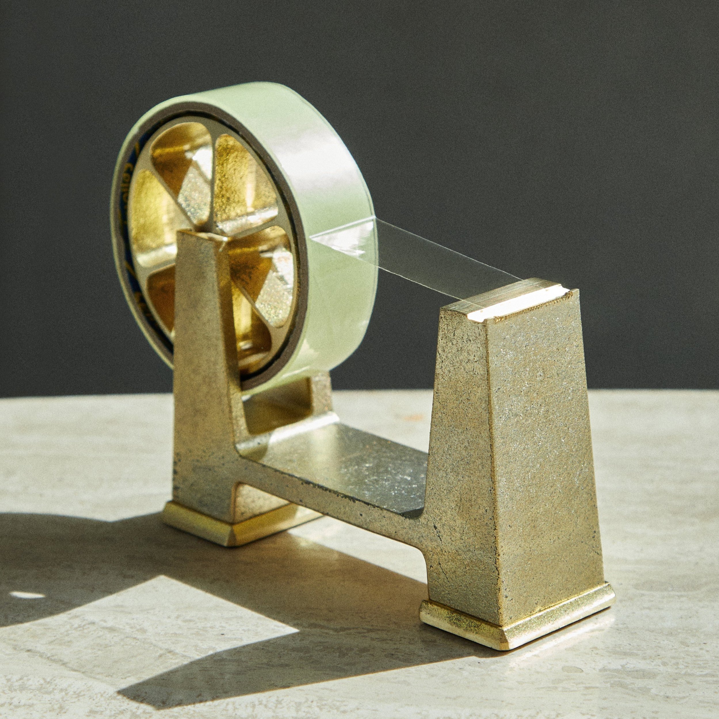 A vintage gold-bound measure tape on a metallic stand with a clear measuring tape extending from it, cast in sunlight on a light-colored surface.