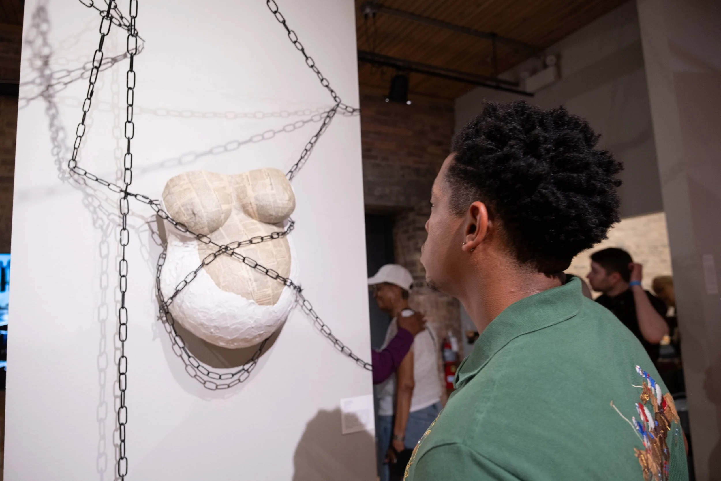 A person with short, curly hair wearing a green shirt is looking at an artwork of a bear's face made from paper and surrounded by black chains on a white background. Other visitors are visible in the background.