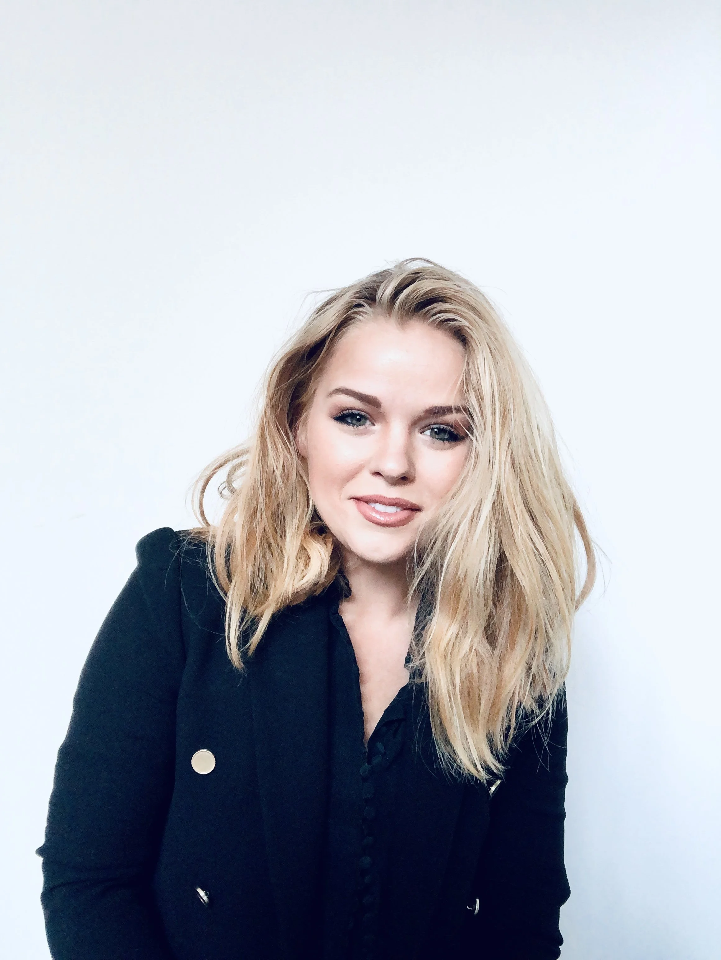 Young woman with blonde hair and light makeup smiling, wearing a black blazer, posed against a plain white background.