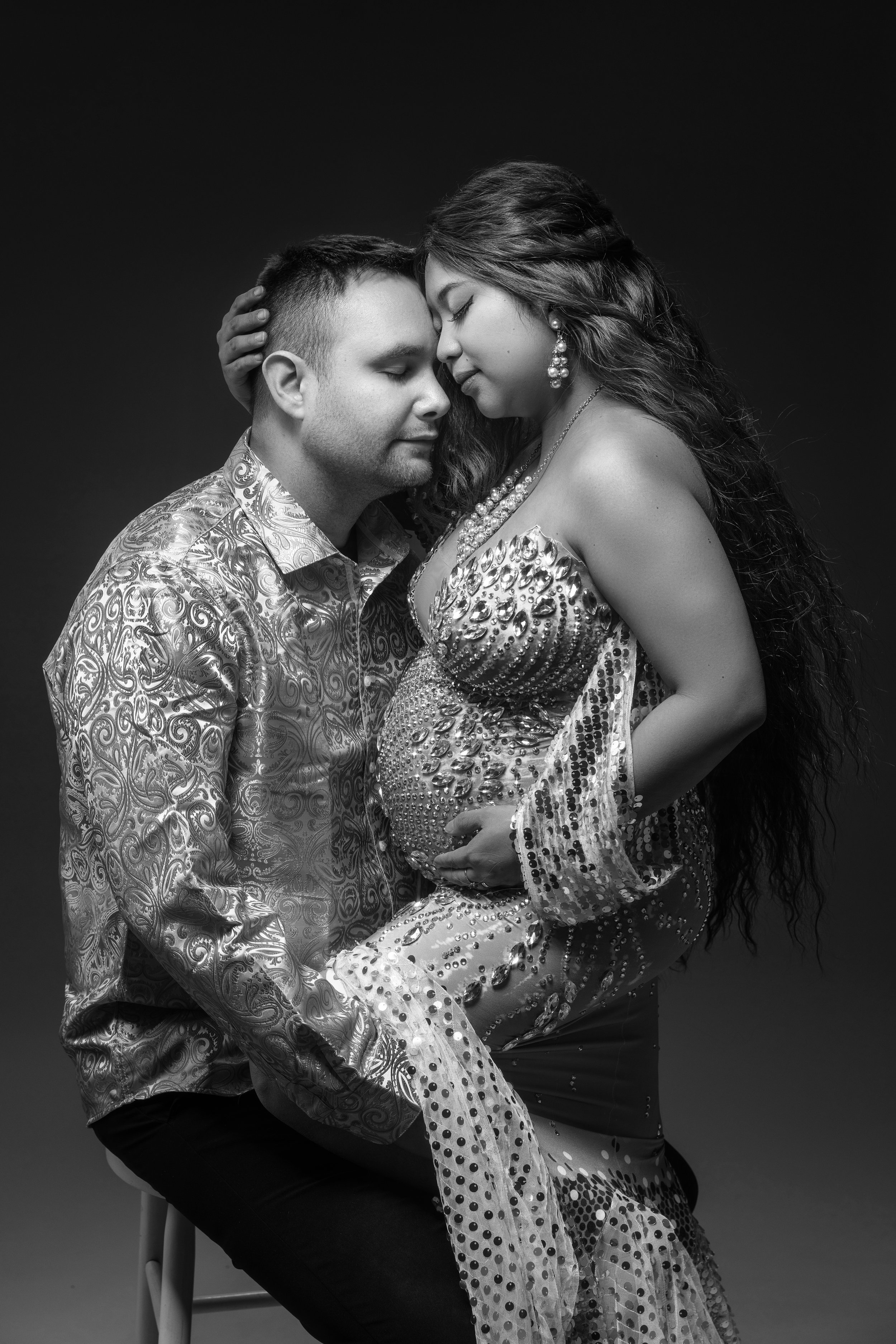 A fashionable black-and-white maternity photo of couple with their foreheads touching, sitting close together against a dark background during their maternity session. Photographed by The Photician.
