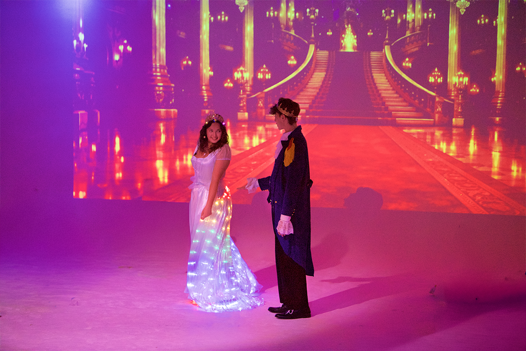 Two people dressed as princess and prince standing on stage with a castle backdrop projection.