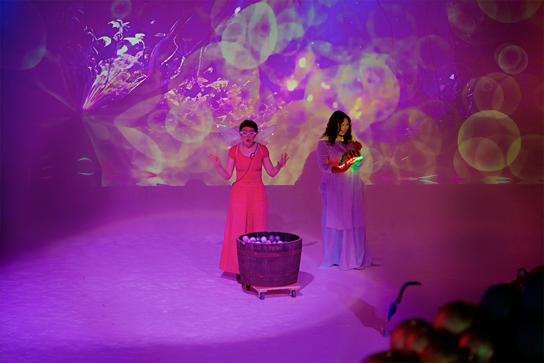 Two women performing on stage with colorful, abstract lighting in the background. One woman is in pink and appears to be talking, while the other woman is in a light-colored dress with glowing objects in her hands. There is a large basket filled with balls in front of them.