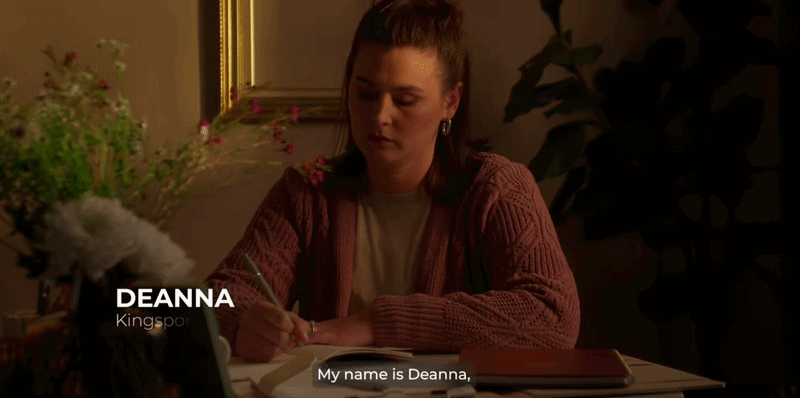 A young woman with brown hair sits at a table writing in a notebook, with a plant and a framed mirror behind her. She is wearing a pink cardigan and appears to be in a cozy indoor setting.