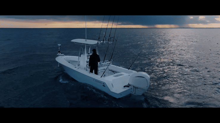 A person standing on a white fishing boat with multiple fishing rods, sailing on the ocean during sunset or sunrise.