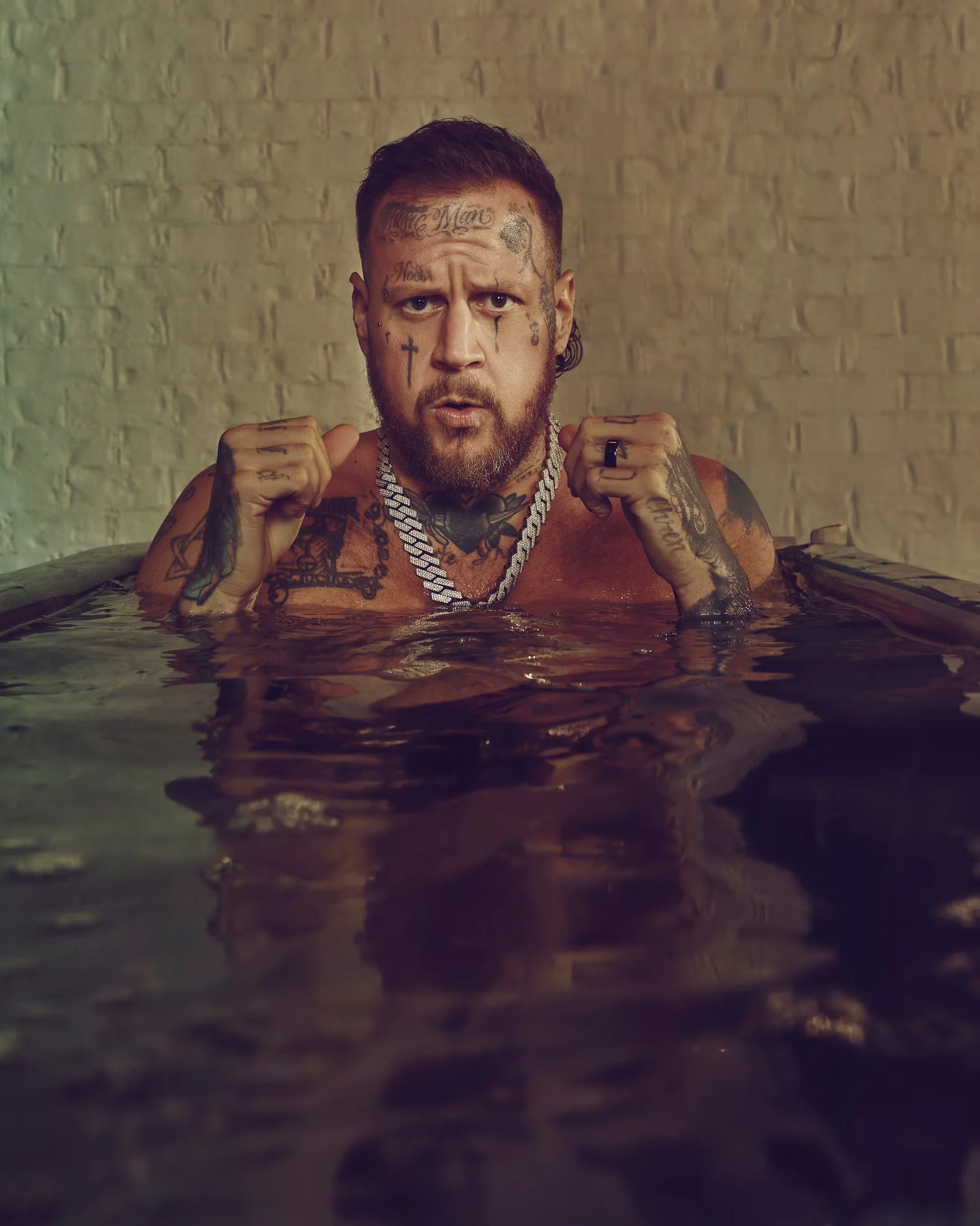 A man with tattoos and jewelry submerged in a dark pool, posing with fists raised, in front of a light-colored brick wall.