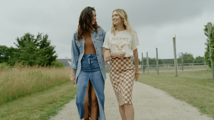 Two women walking on a gravel path outdoors, smiling and looking at each other.