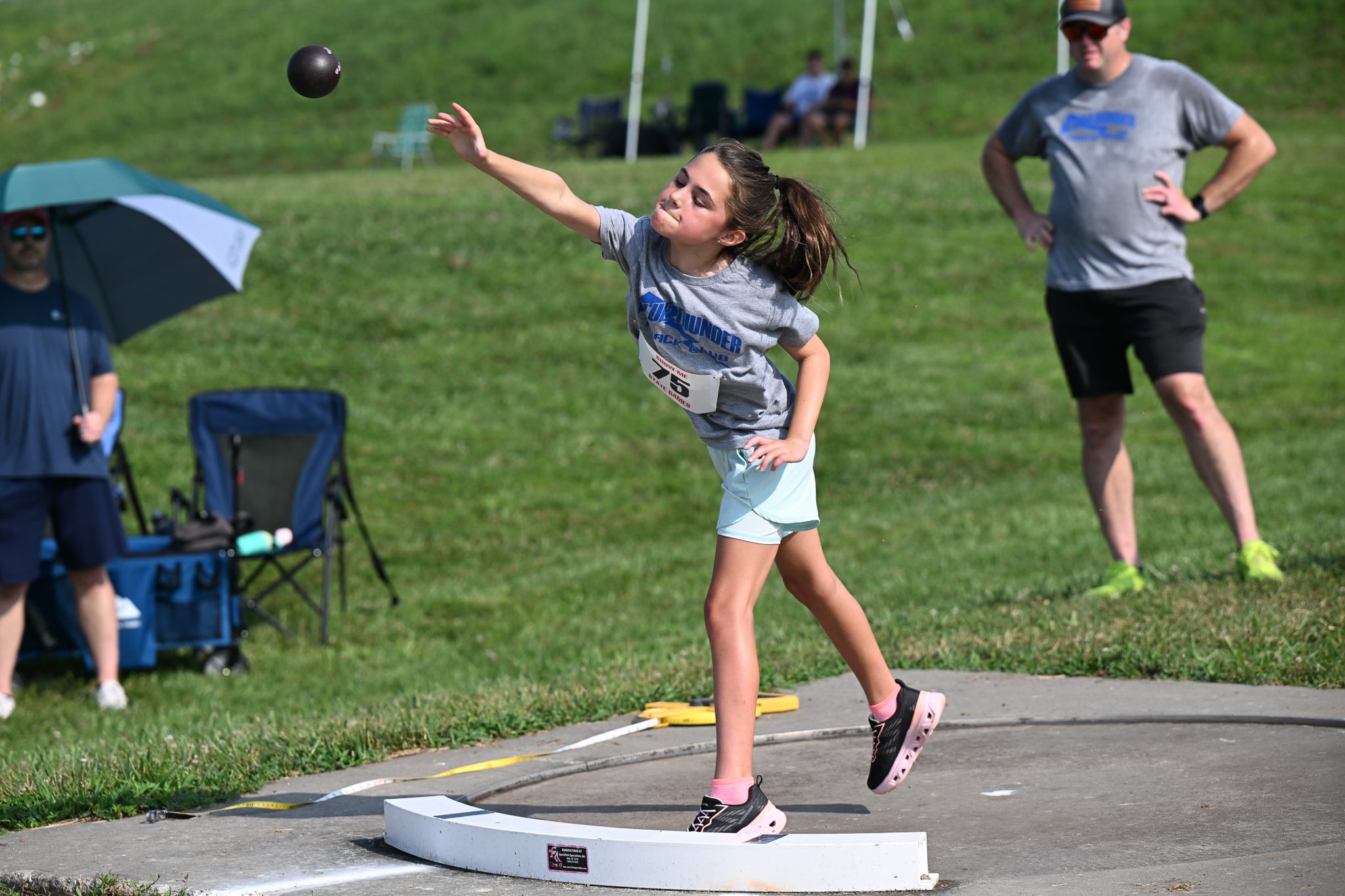 Nora Casey, 9, from Columbia, throws her first attempt in the during the girls shot put as her father, Nathan Casey, coaches from behind during the Show Me State games Track and Field competition on Saturday, July 25, 2026, at Battle High School in C