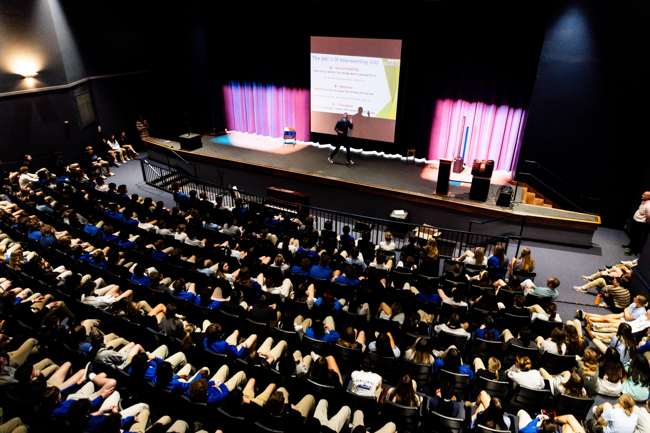 A large auditorium filled with students watching a presentation on stage. A speaker stands in front of a large screen displaying a slide titled 'The ABC's of Representing YOU'. The stage has purple curtains, and there are chairs and equipment set up.