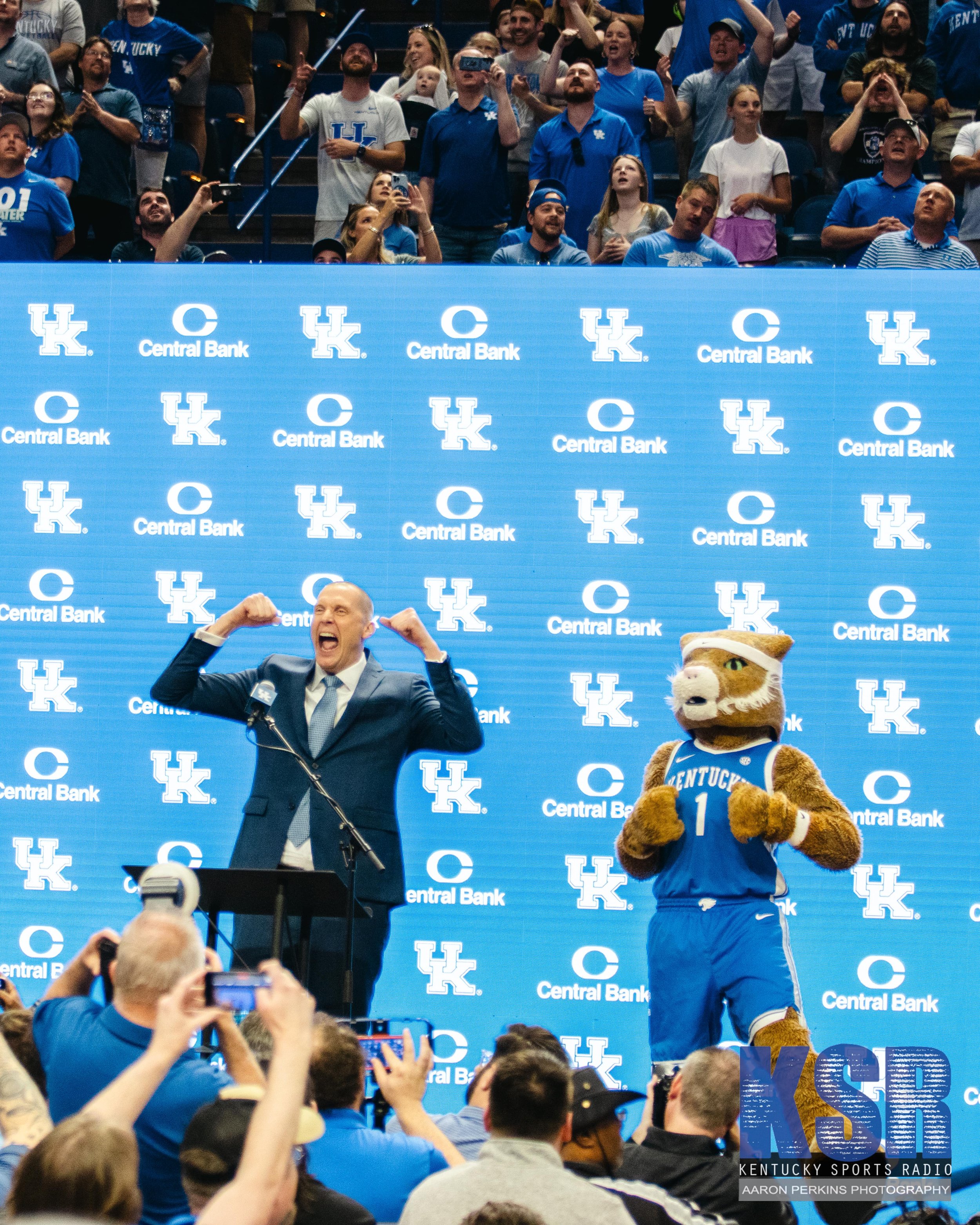 A man in a suit and tie, standing at a podium, is celebrating with arms flexed, and an animal mascot dressed in a Kentucky basketball uniform stands next to him on stage. The background features a blue backdrop with logos for Kentucky and Central Ban