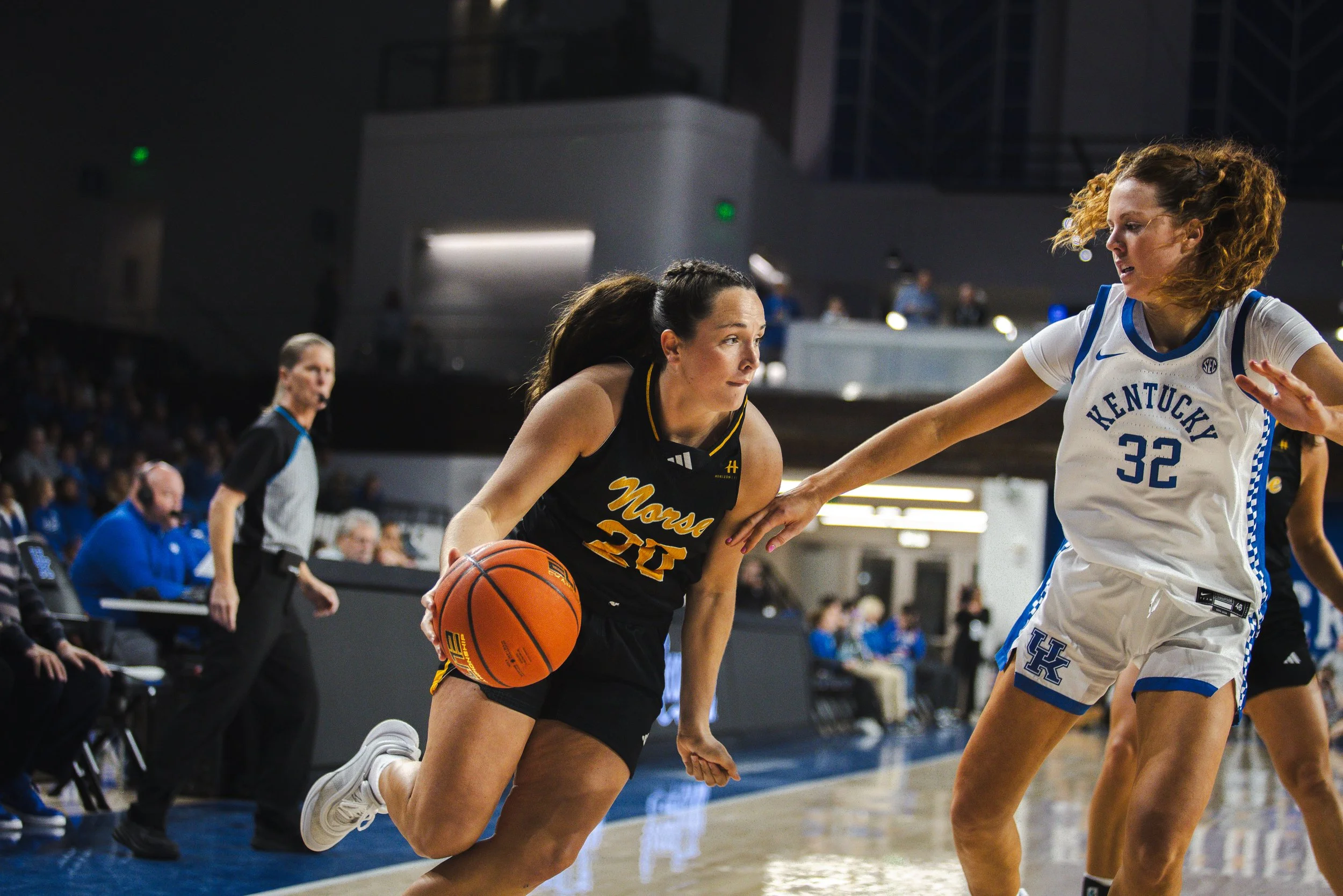 A women's college basketball game between teams from NKU and Kentucky, with a player from Mars dribbling the ball while being guarded by a player from Kentucky.